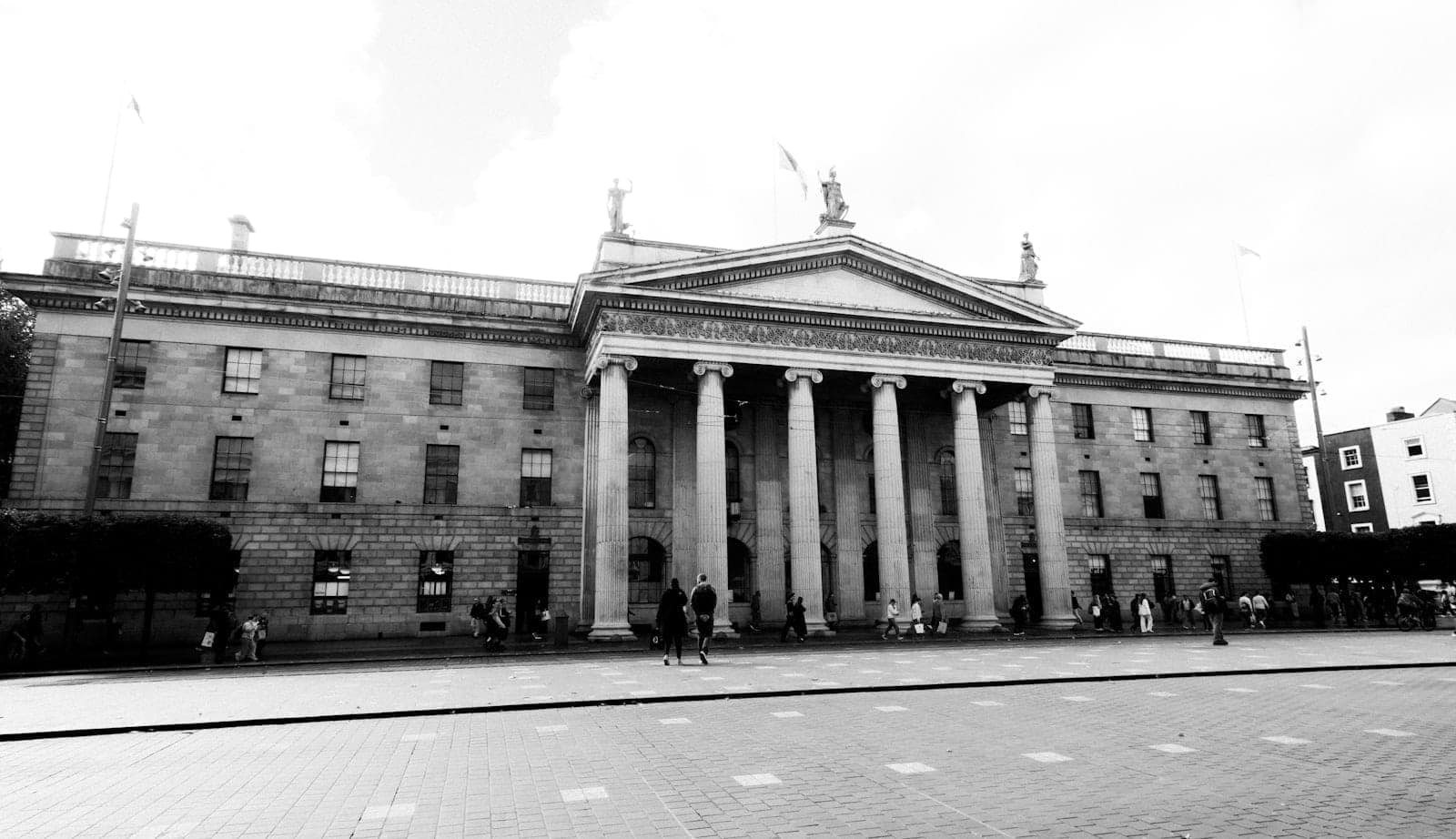 Szerokie czarno-białe zdjęcie Głównego Urzędu Pocztowego w Dublinie z charakterystycznymi kolumnami i przechodniami na O'Connell Street.