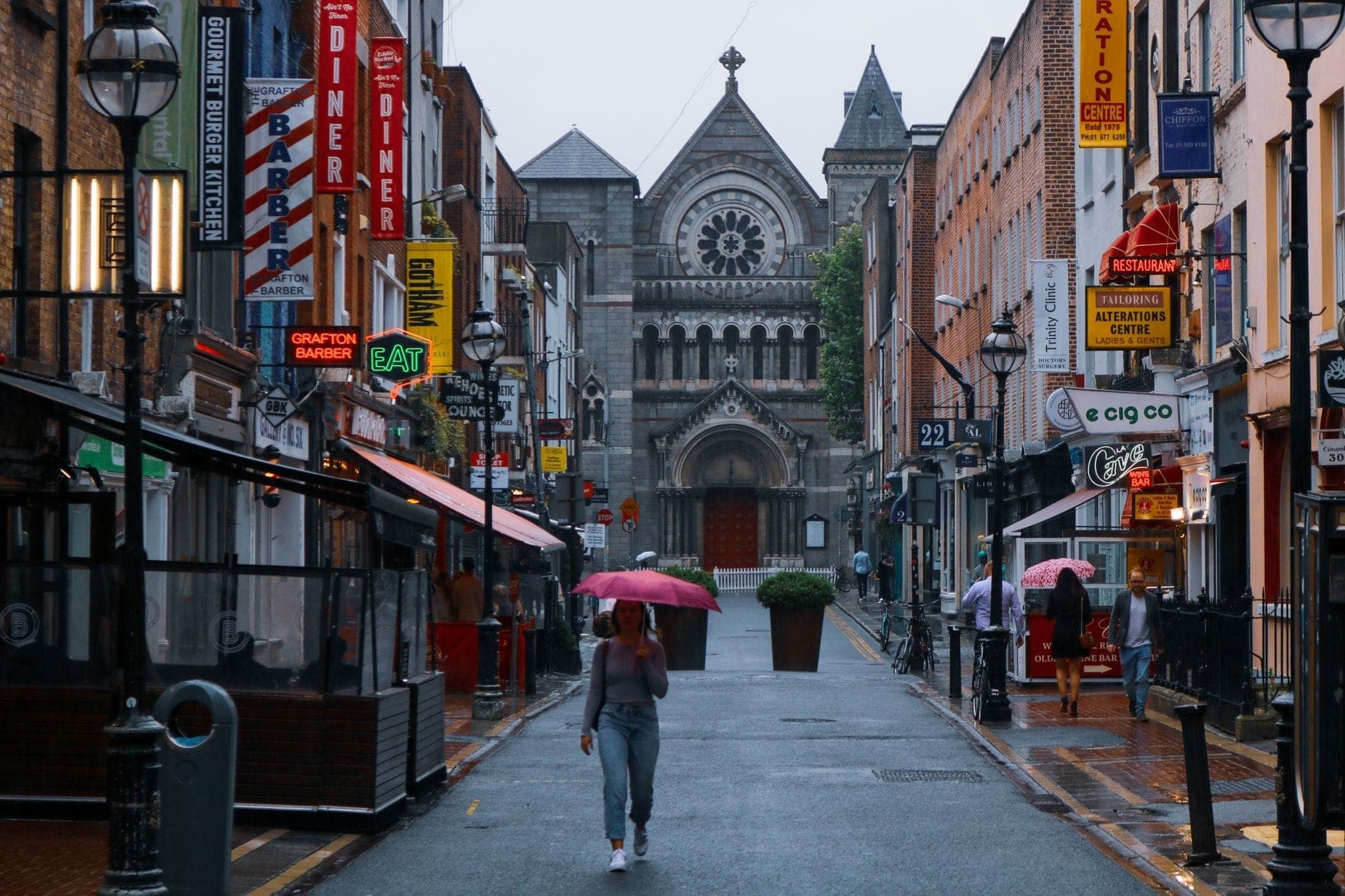 Vue de Grafton Street à Dublin un jour de pluie, avec des enseignes de magasins colorées, des piétons et une église historique au bout de la rue.
