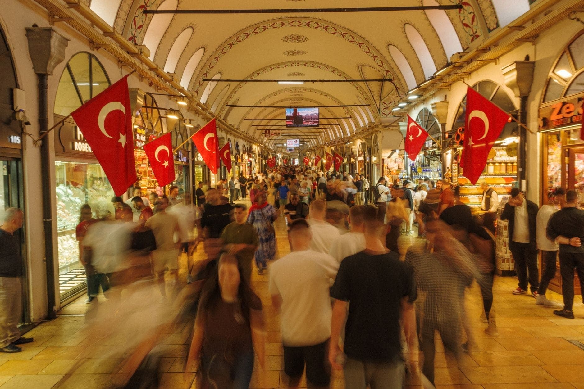 Belebtes Inneres des Großen Basars in Istanbul: Menschenmassen, türkische Fahnen, gewölbte Decken und bunte Geschäfte auf beiden Seiten eines breiten Korridors.