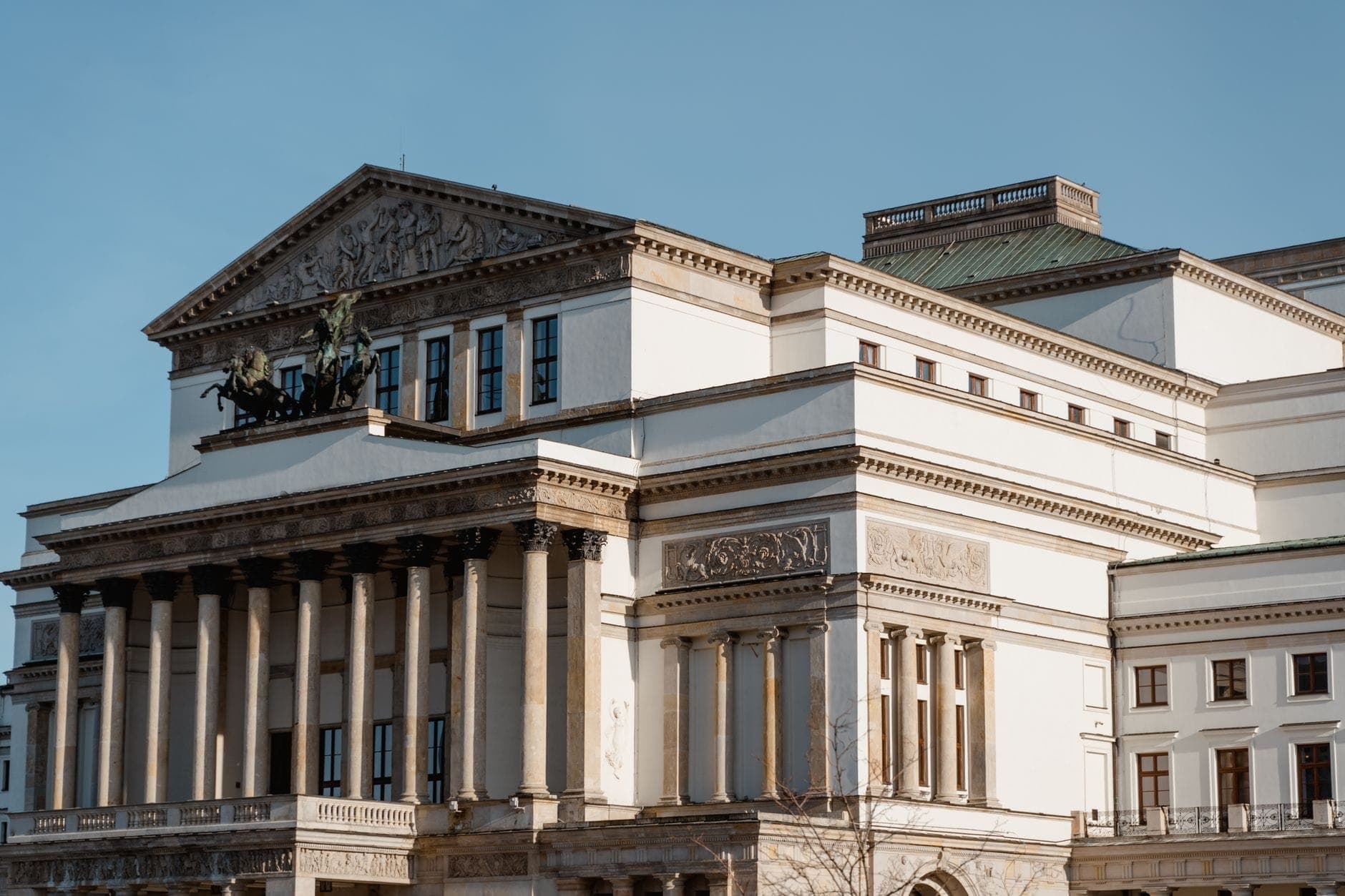 Grand Theatre – Nationaloper Warschau mit einer neoklassischen Fassade mit hohen Säulen, kunstvollen Schnitzereien und einer bronzenen Wagenplastik vor klarem blauem Himmel.