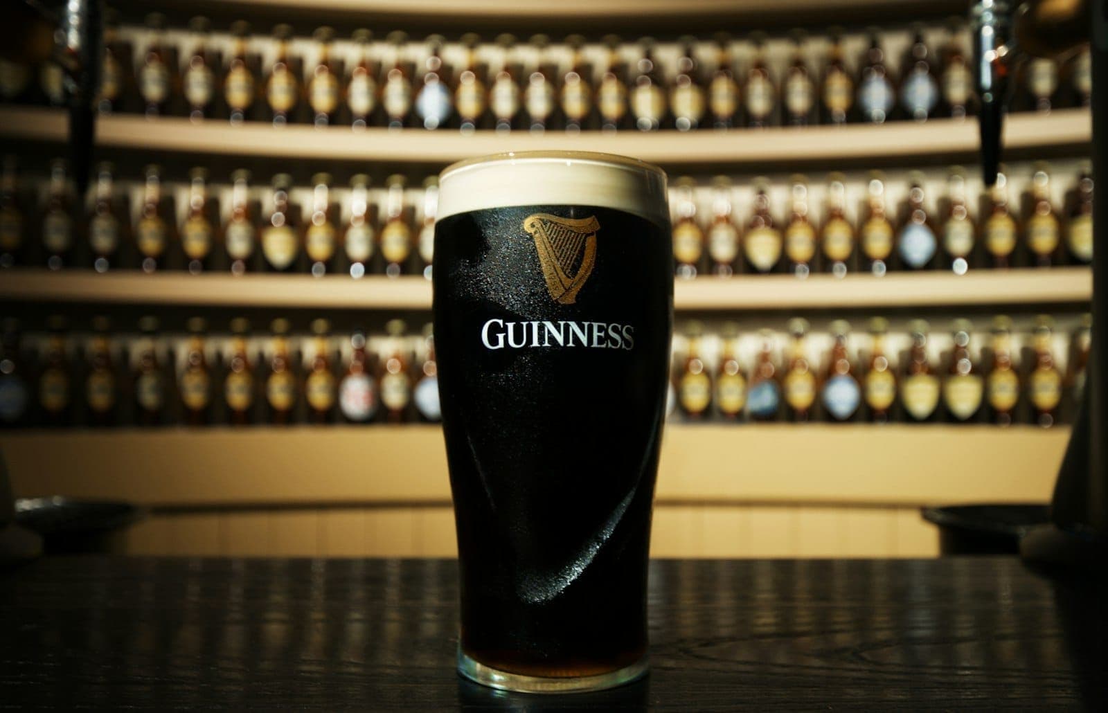 Una pinta di Guinness spillata alla perfezione riposa su un bancone lucido, con decine di bottiglie di birra diverse allineate lungo la parete curva dietro, in un taproom dall'illuminazione calda.