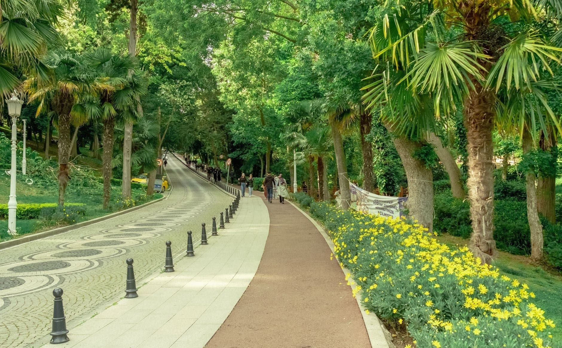 Geschwungene Wege gesäumt von gelben Blumen, Palmen und Besuchern, die an einem sonnigen Tag durch das üppige Grün des Gülhane Parks schlendern.
