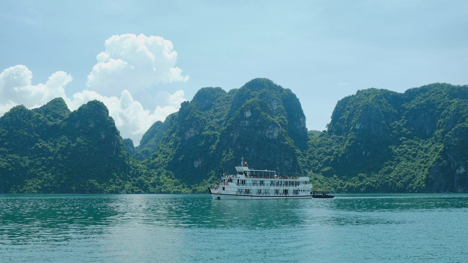 Một chiếc du thuyền trắng lớn lướt qua làn nước xanh ngọc bích, xung quanh là những núi đá vôi hùng vĩ dưới bầu trời xanh trong tại Vịnh Hạ Long, Việt Nam.