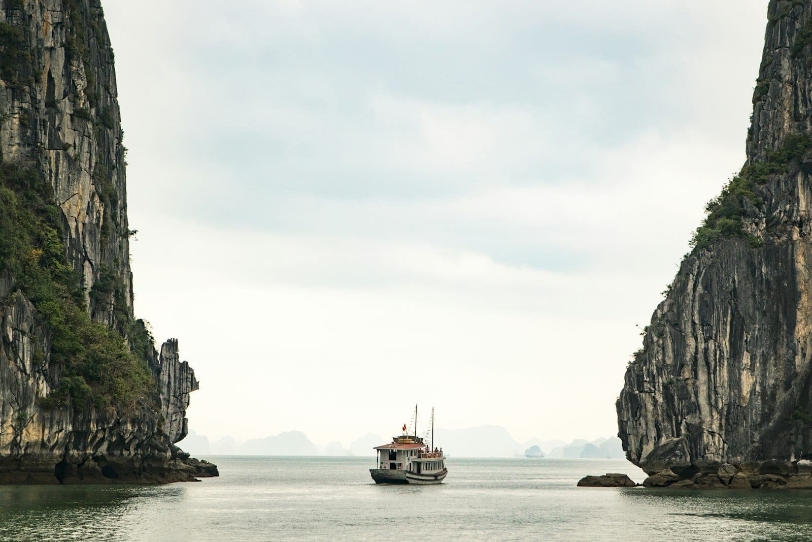 Một du thuyền lướt giữa những vách đá vôi hùng vĩ ở Vịnh Hạ Long dưới bầu trời nhiều mây, khung cảnh biển đảo ấn tượng với những khối karst đặc trưng.