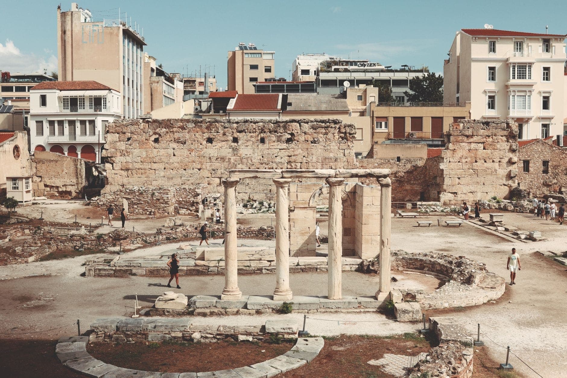 Wide view of the ruined columns and foundations of Hadrian's Library with ancient stone walls and surrounding modern Athens buildings under a clear sky.