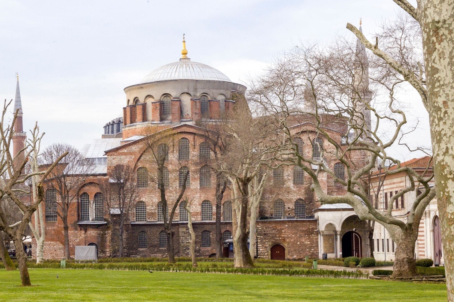 Weitwinkelpanorama der Außenfassade des Hagia-Irene-Museums mit der markanten Kuppel, rohem Backsteinmauerwerk, Bogenfenstern und kahlen Bäumen in den Gärten des Topkapı-Palastes.