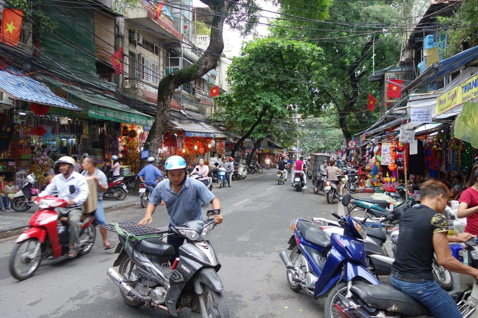 Phố Cổ Hà Nội nhộn nhịp với xe máy, hàng cây, cửa hàng và người qua lại — cảnh sống động thường ngày ở thủ đô Việt Nam.