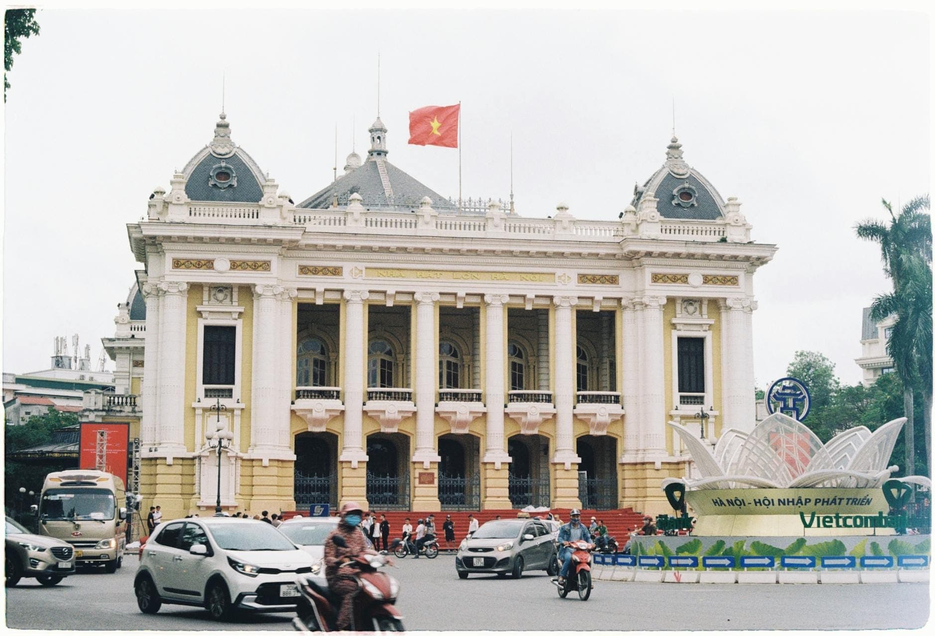 Nhà hát Lớn Hà Nội chụp ban ngày với lá cờ Việt Nam trên đỉnh, toàn cảnh mặt tiền tân cổ điển, dòng xe cộ tấp nập và người qua lại phía trước.