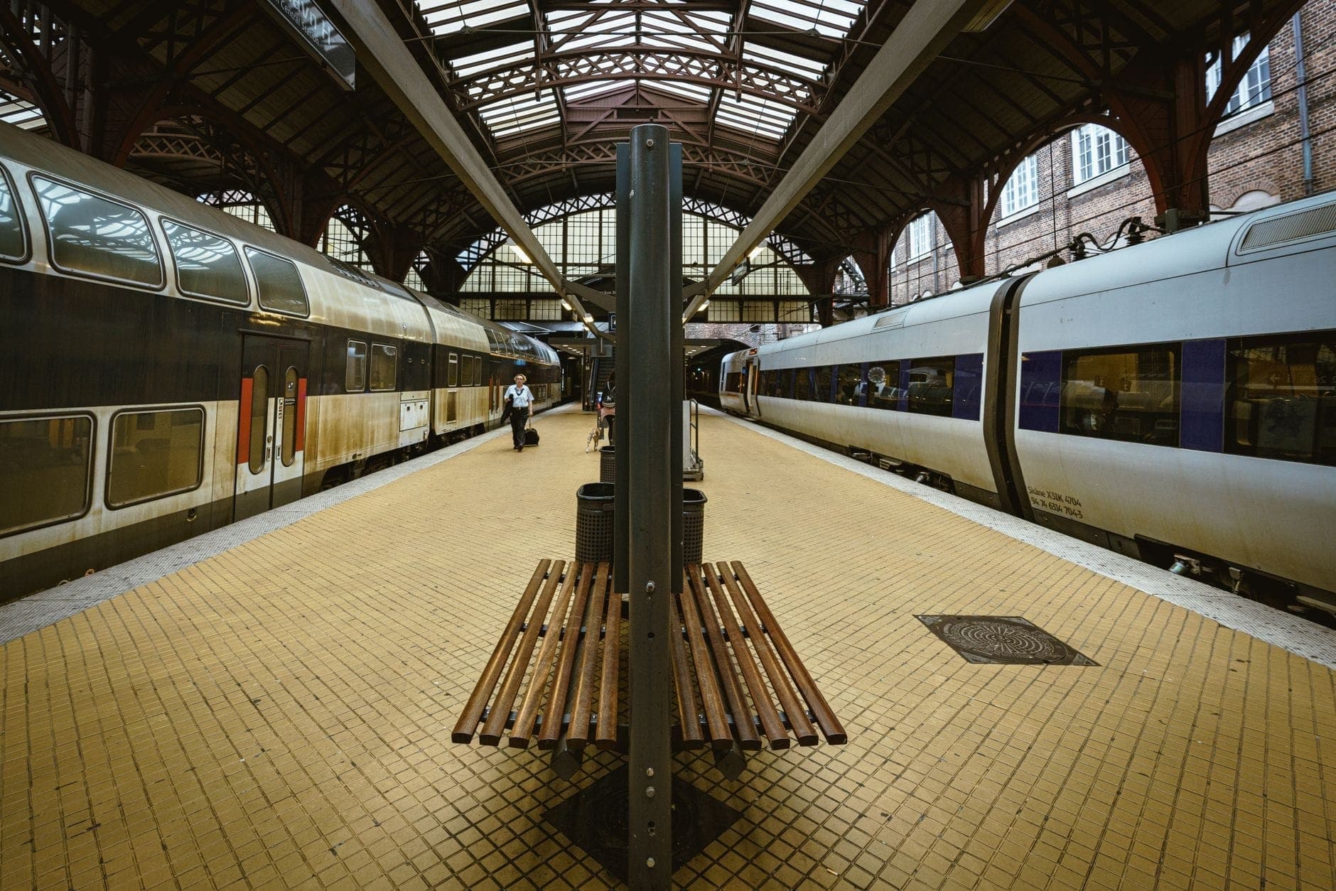 Binario della Stazione Centrale di Copenaghen con panchine, passeggeri e tipici treni danesi sotto un grande tetto ad arco.