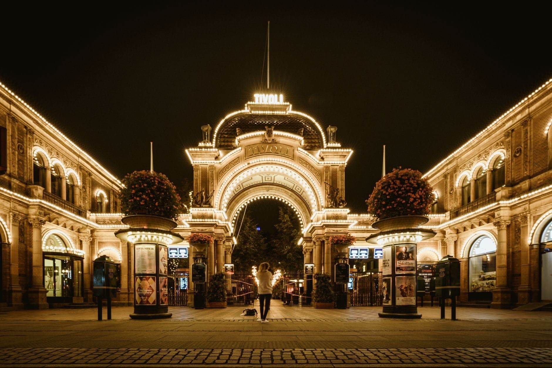 L'arco illuminato dell'ingresso principale dei Tivoli Gardens di notte, con luci brillanti che delineano l'architettura storica e una persona in piedi al centro.