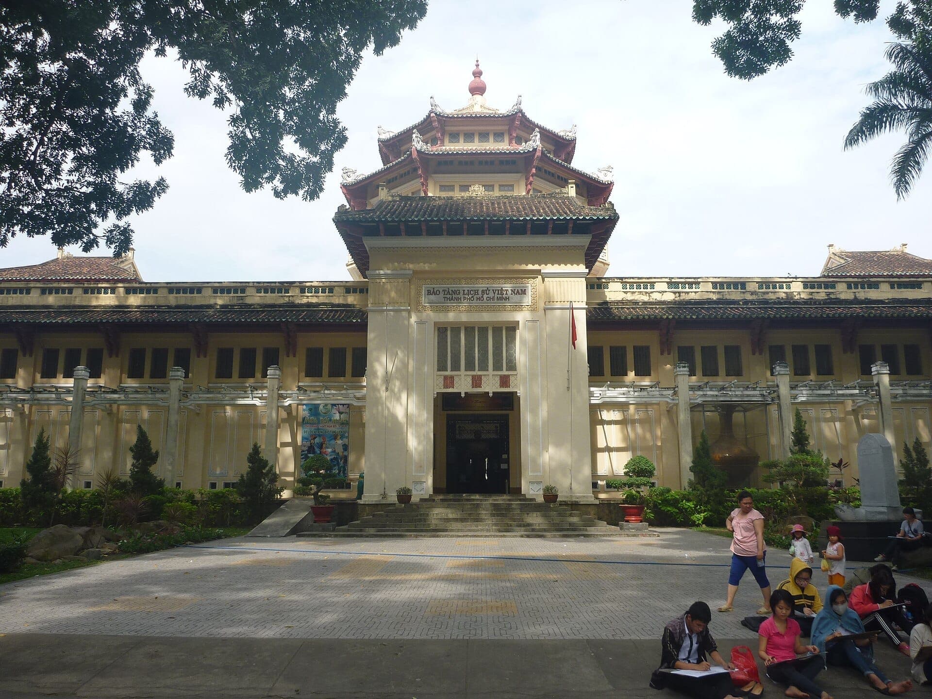 Mặt tiền Bảo tàng Lịch sử Thành phố Hồ Chí Minh với kiến trúc thuộc địa, đường mái kiểu chùa và du khách ngồi nghỉ trong sân tiền sảnh có bóng mát.