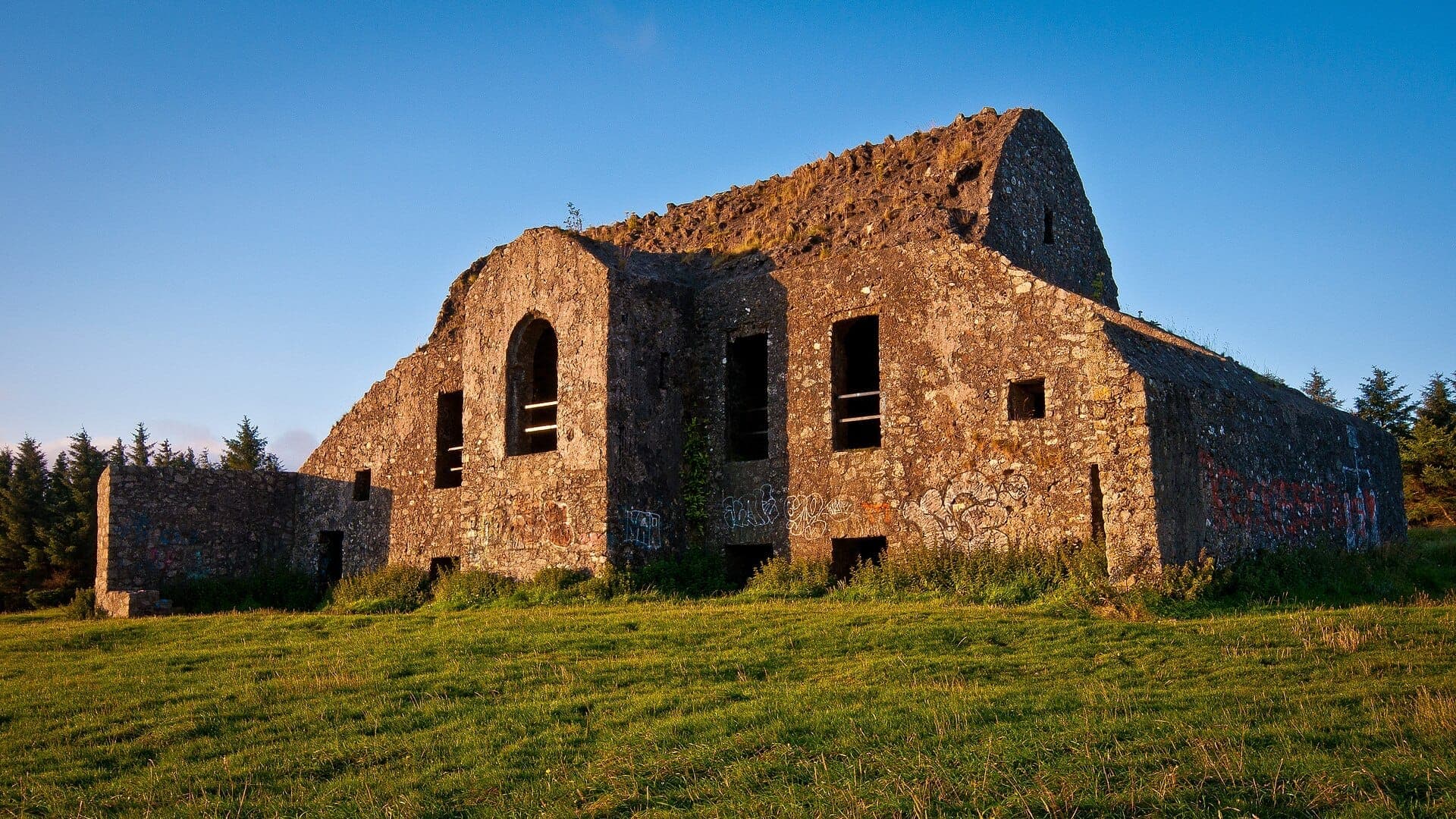 Il rudere dell'Hellfire Club si trova sul verde Montpelier Hill sotto un cielo azzurro limpido, circondato da alberi sempreverdi lontani e dalla luce dorata della sera.