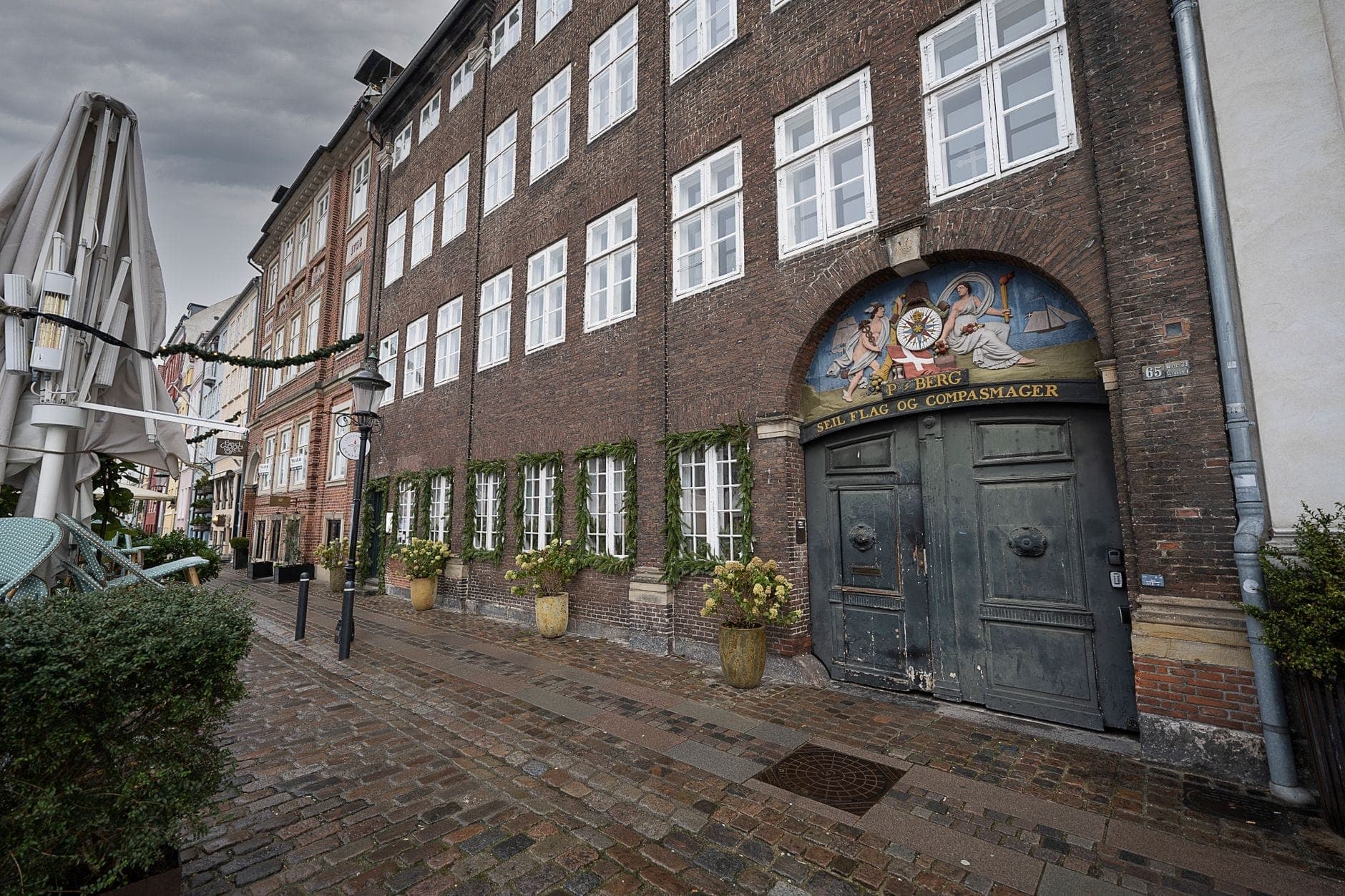 Eine Kopfsteinpflasterstraße in Kopenhagen, gesäumt von alten Backsteingebäuden, großen Fenstern und grünen Pflanzenkübeln, unter einem bewölkten Himmel ohne Menschenmassen.