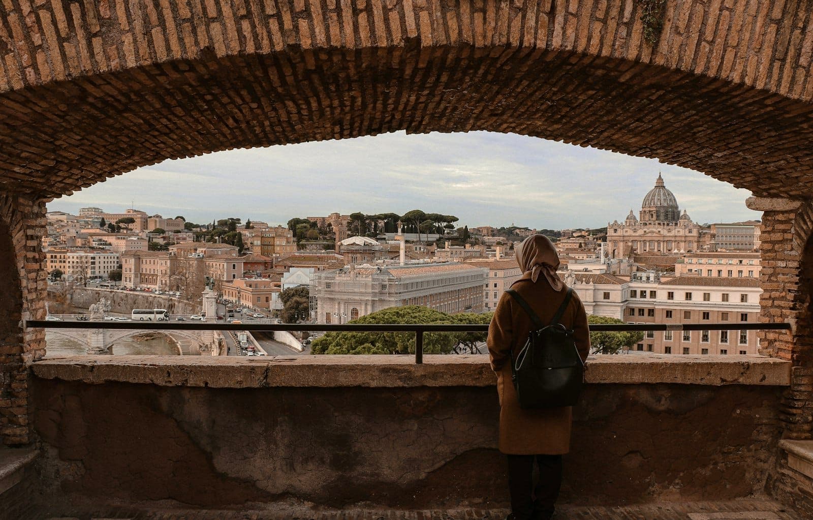 Một người đeo ba lô nhìn ra mái nhà của Rome qua một vòm đá, với Vương cung thánh đường Thánh Phêrô hiện ra ở phía xa.