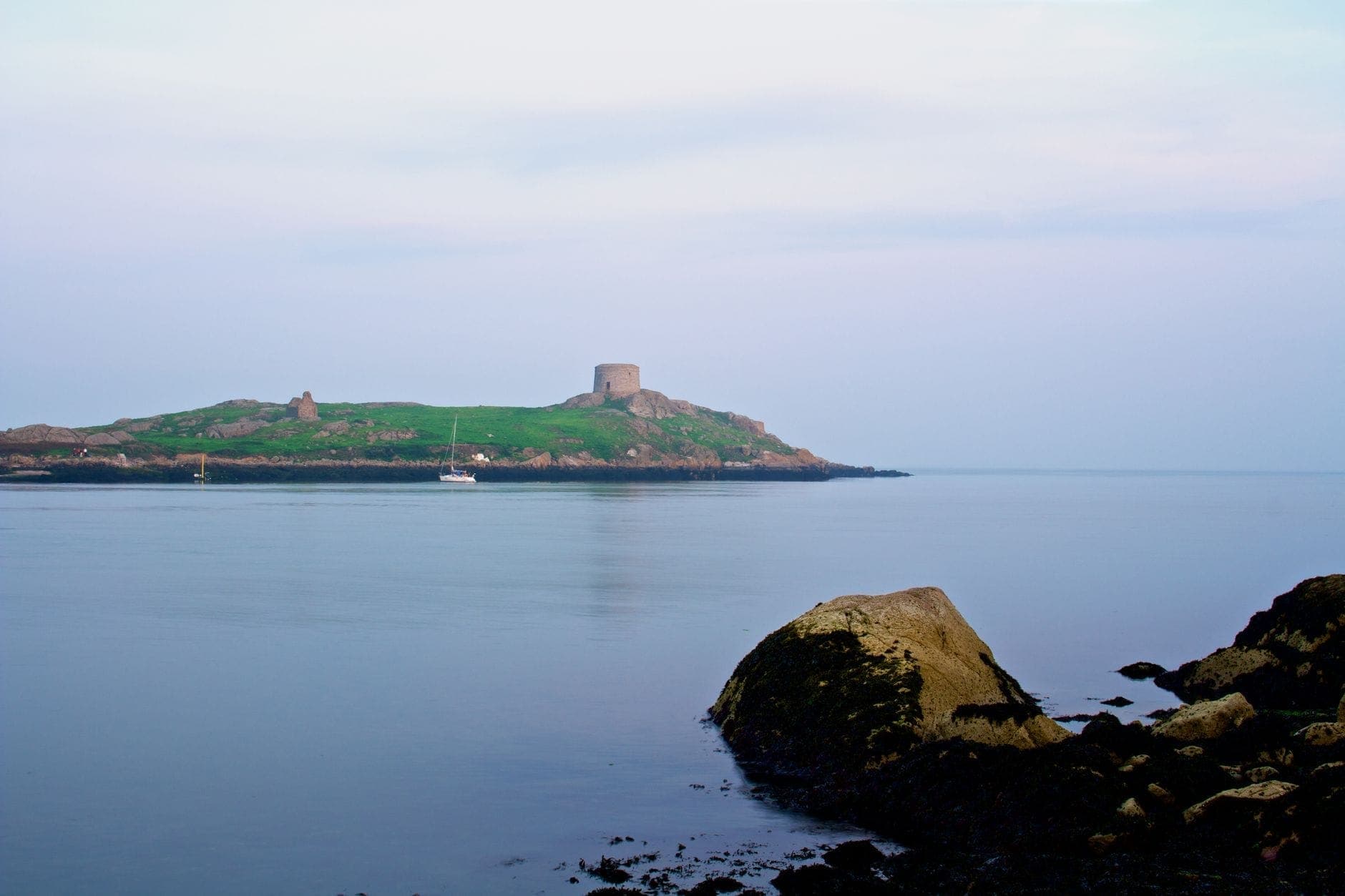 Vista lontana di Dalkey Island con la sua torre Martello, vista dalla riva con mare calmo e rocce in primo piano.