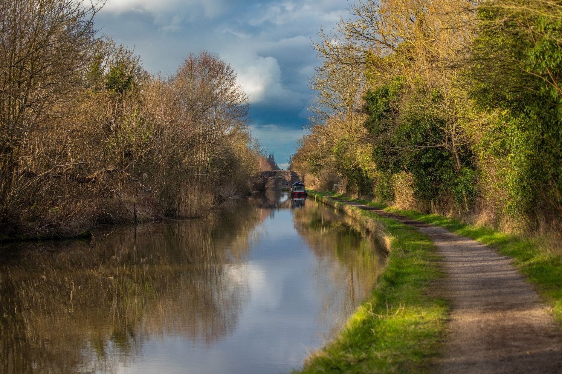 Un tranquillo sentiero lungo il canale fiancheggiato da rive erbose e alberi con una narrowboat e un ponte in lontananza, ideale per passeggiate nella natura vicino a Dublino.
