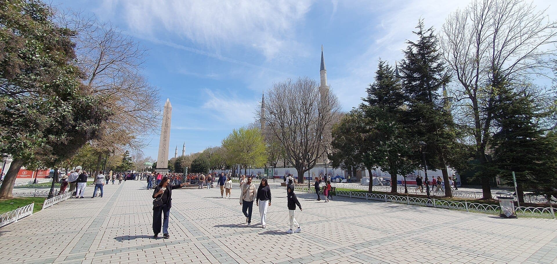Weitläufiger Steinplatz am Hippodrom von Konstantinopel, mit dem antiken Ägyptischen Obelisken des Theodosius, Minaretten, Bäumen und Menschen, die unter einem blauen Himmel spazieren.