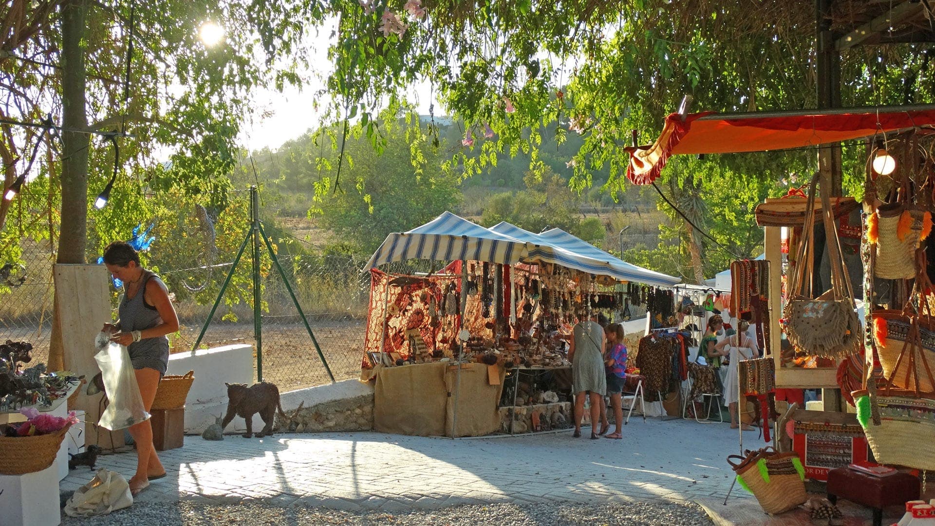 Outdoor stalls at Las Dalias Hippy Market in Ibiza display colorful handmade bags, jewelry, and crafts under striped awnings in the afternoon sun.