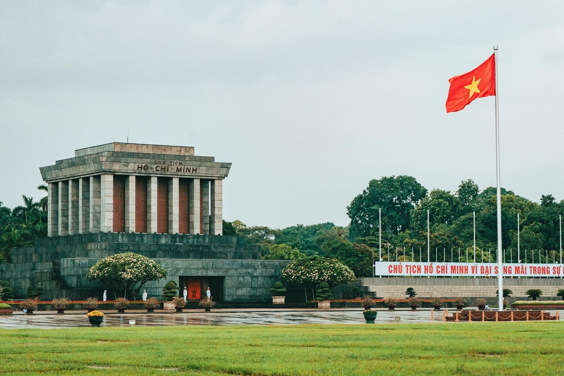 Toàn cảnh Lăng Chủ tịch Hồ Chí Minh tại Hà Nội với cờ Tổ quốc và khuôn viên cây xanh bao quanh trong một ngày trời nhiều mây.