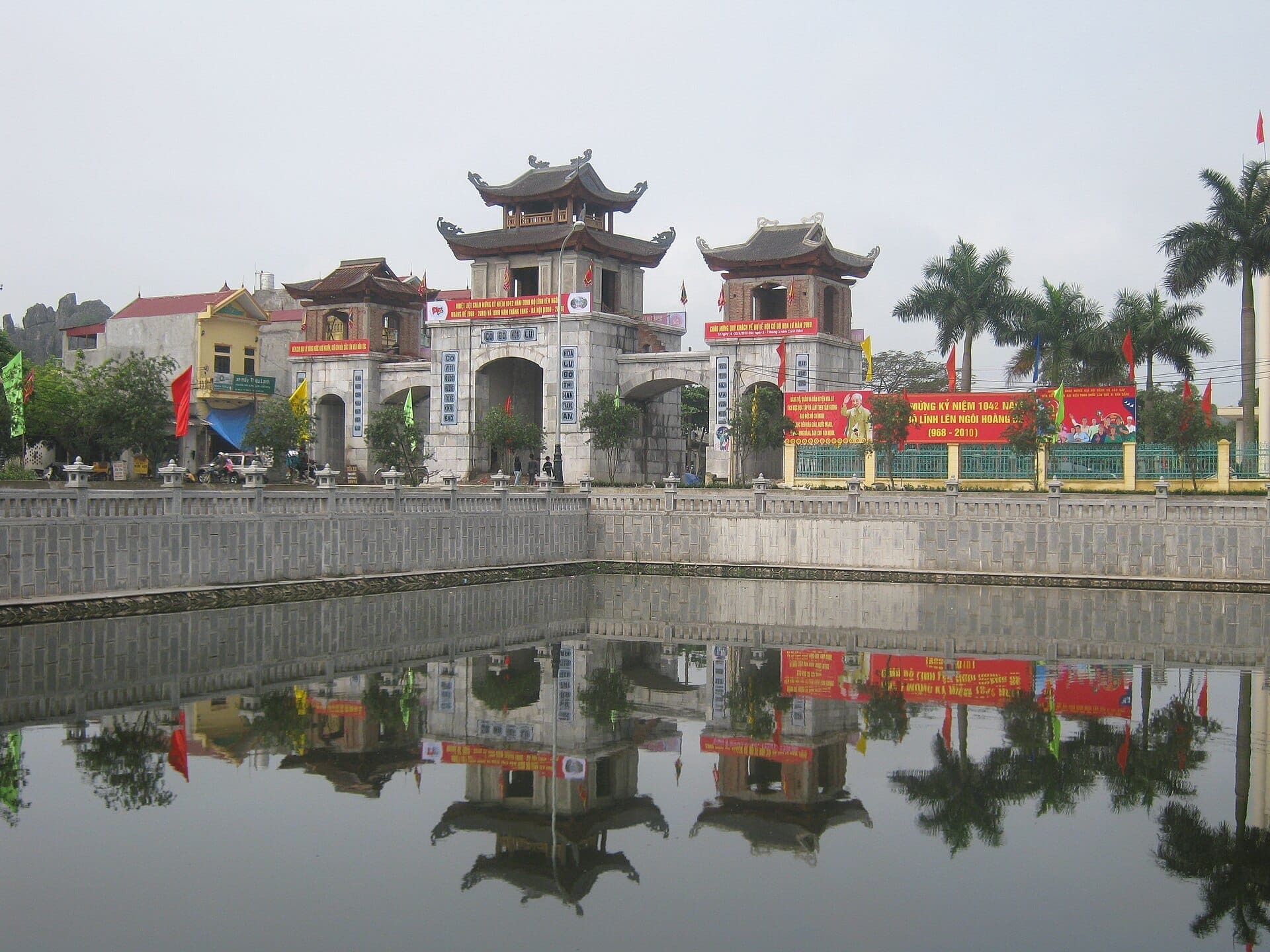 Cổng vào chính của Cố đô Hoa Lư phản chiếu trên mặt hồ yên tĩnh, bao quanh bởi cây cọ, cờ phướn rực rỡ và kiến trúc truyền thống Việt Nam.