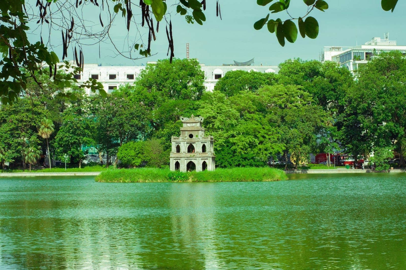 Tanawin ng Hoan Kiem Lake na may Turtle Tower sa maliit na isla, napapalibutan ng mga puno at mga gusali ng lungsod sa likod, nakabalangkas ng mga dahon na sanga.
