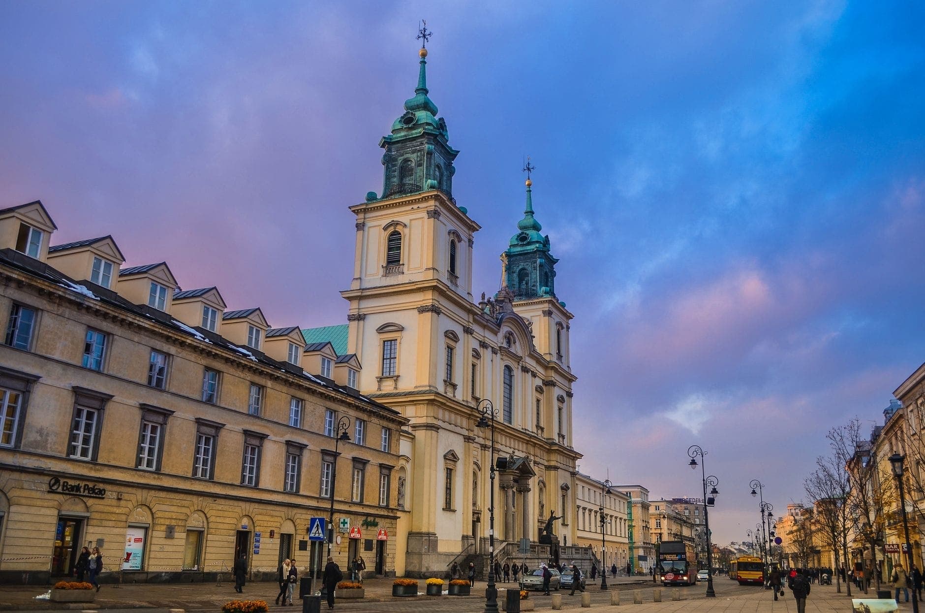 Die Heilig-Kreuz-Kirche in Warschau mit ihren zwei Türmen und der Barockfassade im Sonnenuntergang, eingerahmt von farbenprächtigen Abendhimmel und belebtem Krakowskie Przedmieście.