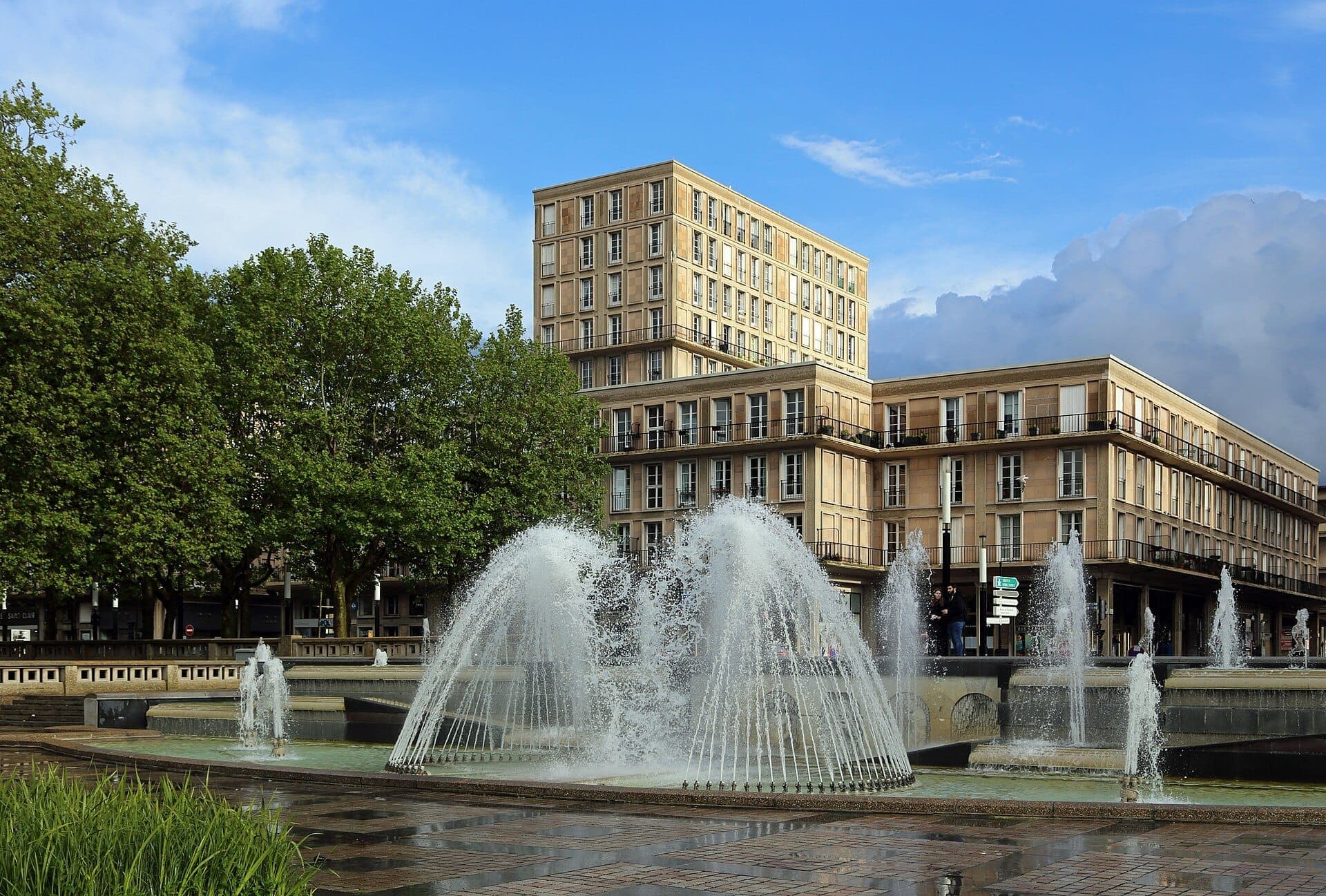 Hôtel de Ville du Havre sừng sững phía sau đài phun nước sôi động và những cây xanh dưới bầu trời xanh trong.