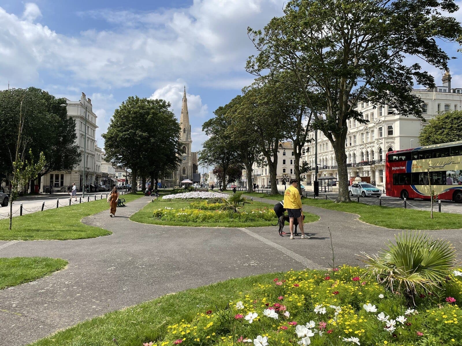 Malawak na boulevard sa Hove na may mga taniman ng bulaklak, malalaking puno, makasaysayang puting gusali, isang kampanario ng simbahan, at isang pulang bus ng Brighton & Hove sa ilalim ng asul na langit.