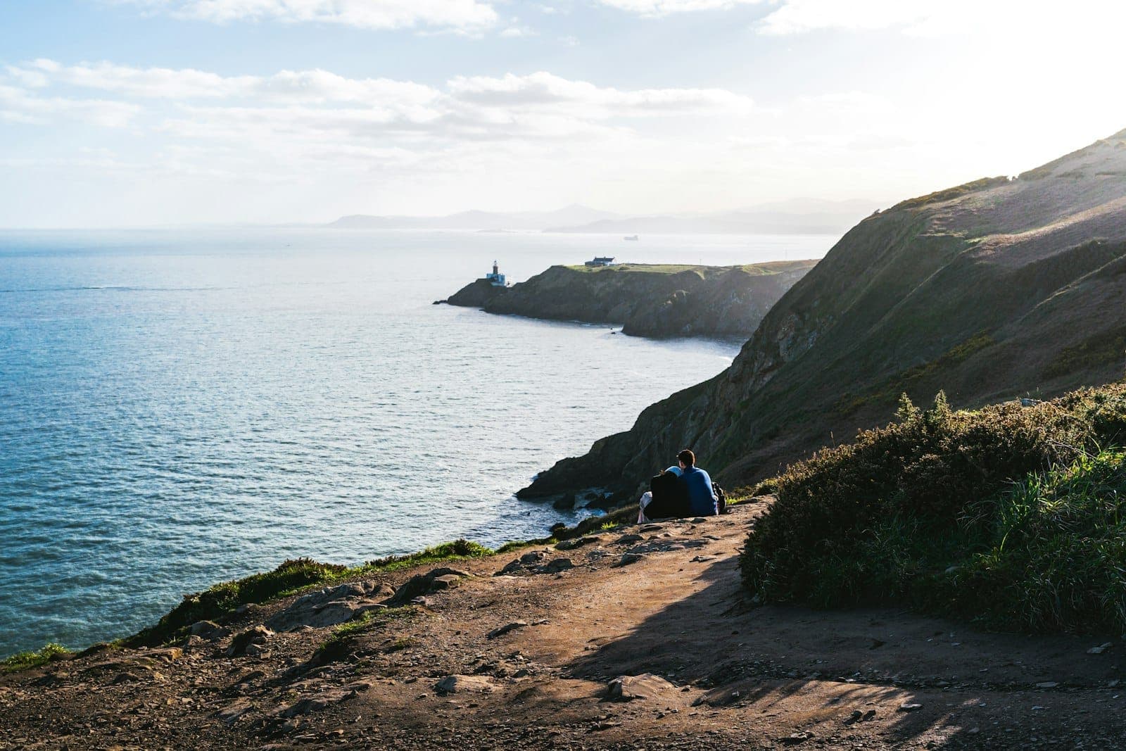 Zwei Menschen sitzen am Howth Cliff Walk und blicken auf dramatische Klippen, die Irische See und einen weit entfernten Leuchtturm unter klarem Himmel.