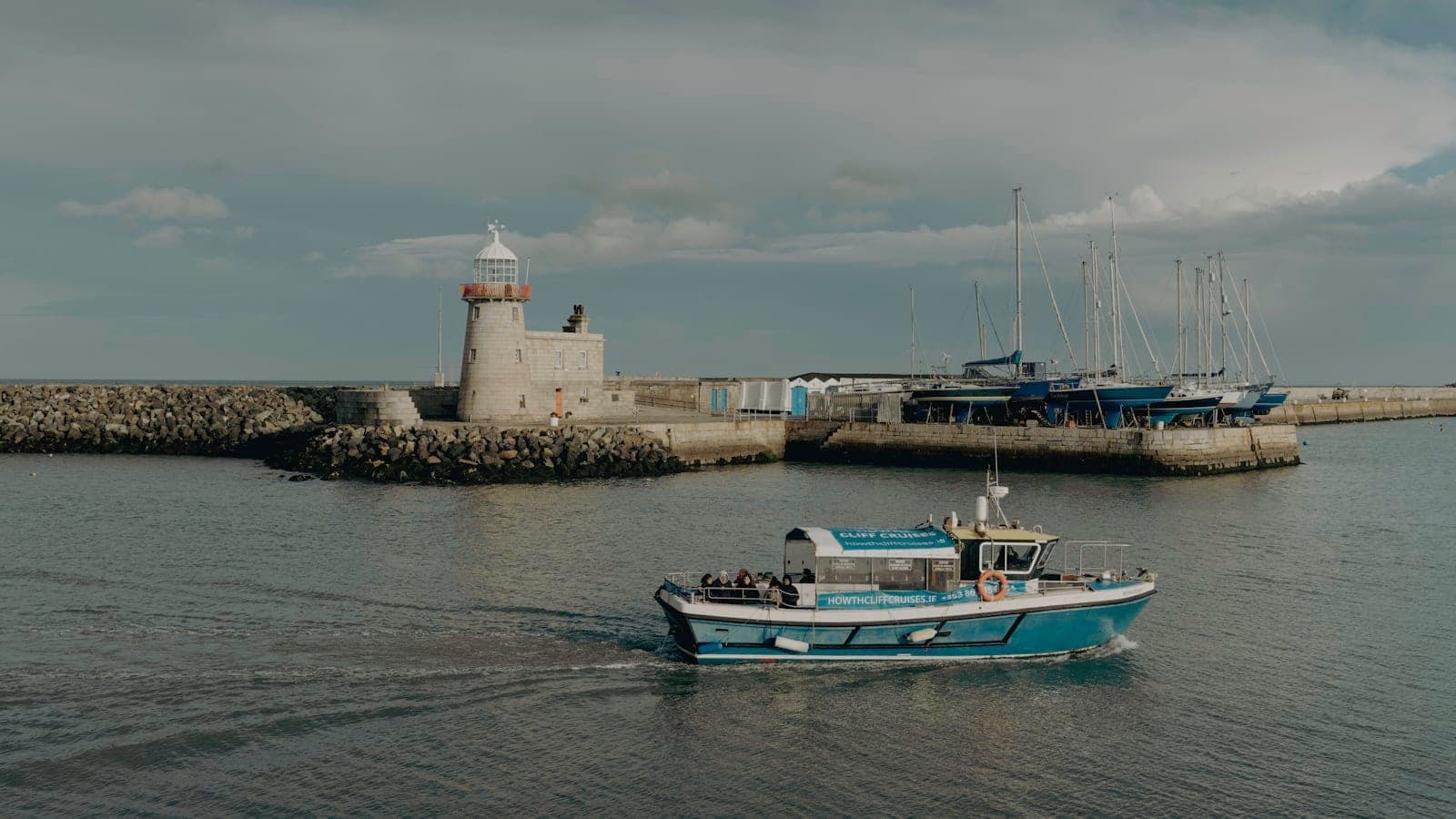Un barco de pesca pasa junto al emblemático faro del puerto de Howth, con veleros y el rompeolas al fondo bajo un cielo luminoso y nublado.