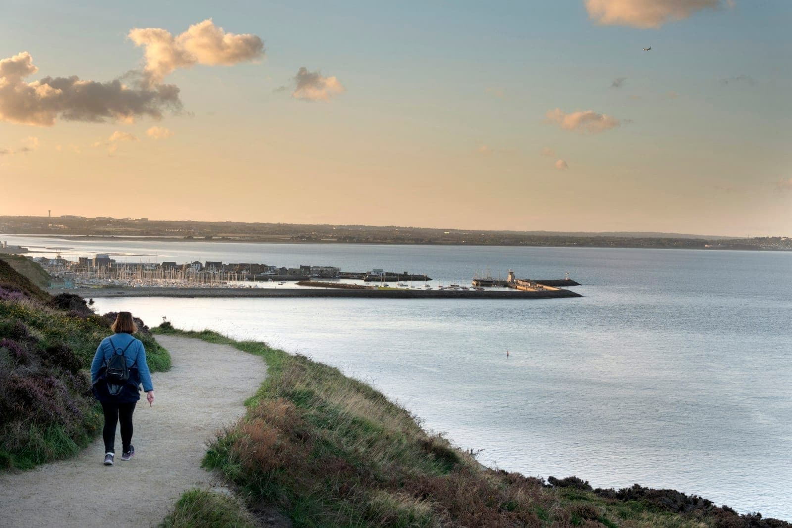 Eine Person wandert auf einem Klippenweg mit Blick auf den Hafen von Howth, die Marina und den Leuchtturm bei Sonnenuntergang – dramatischer Himmel, traumhafte Küstenlandschaft.