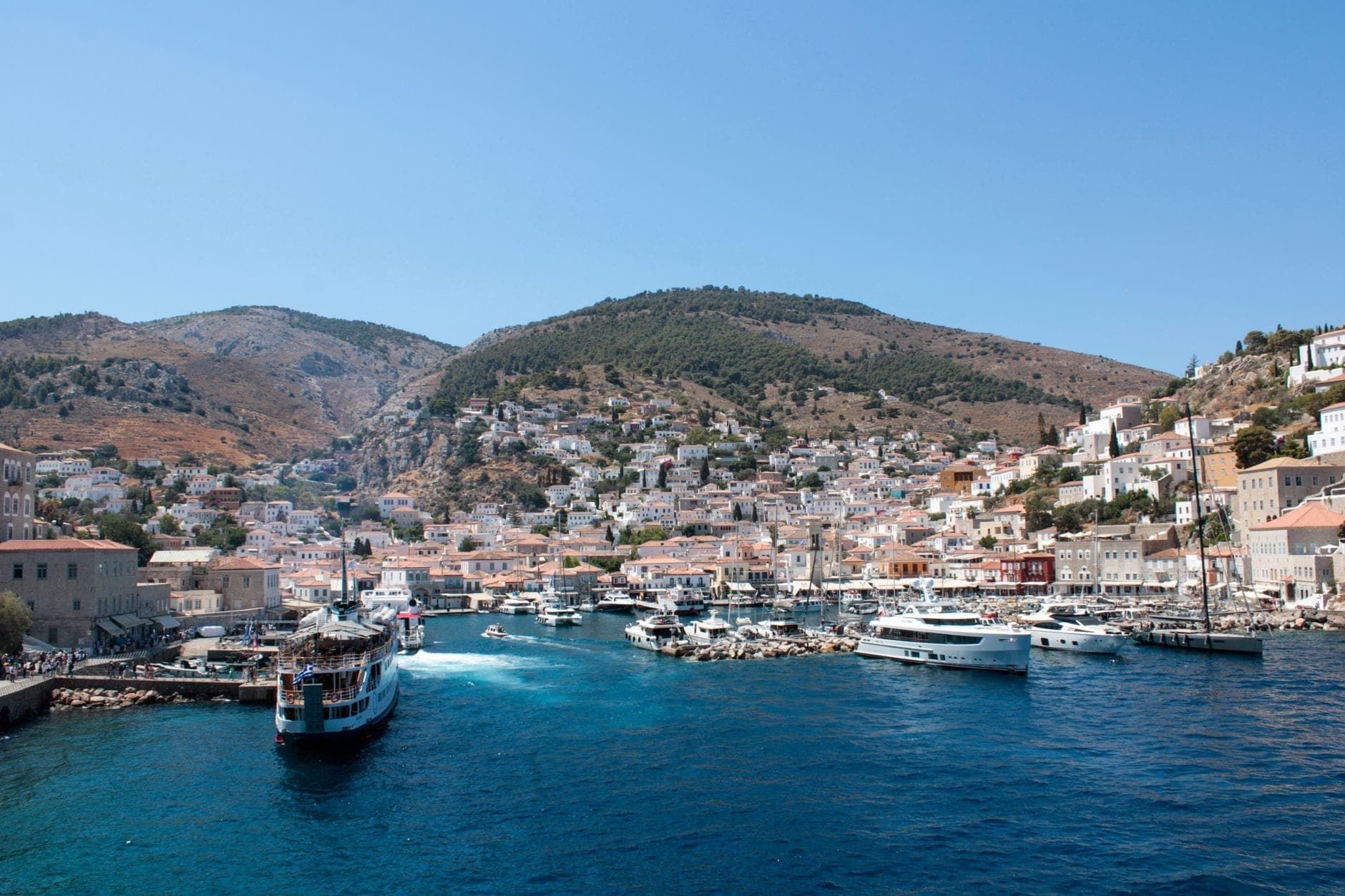 Wide view of Hydra Island’s harbor with boats docked along the waterfront, whitewashed houses climbing the hillside, and rugged mountains in the background under clear blue sky.