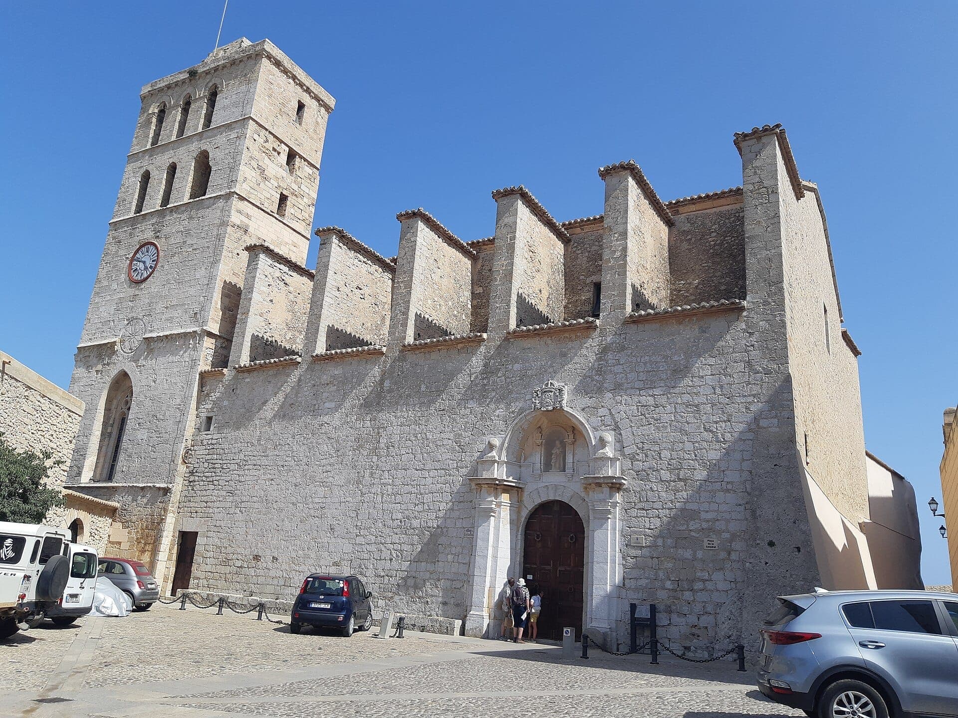 Ibiza Cathedral’s stone facade and clock tower rise against a clear blue sky, with parked cars and visitors near the historic entrance.