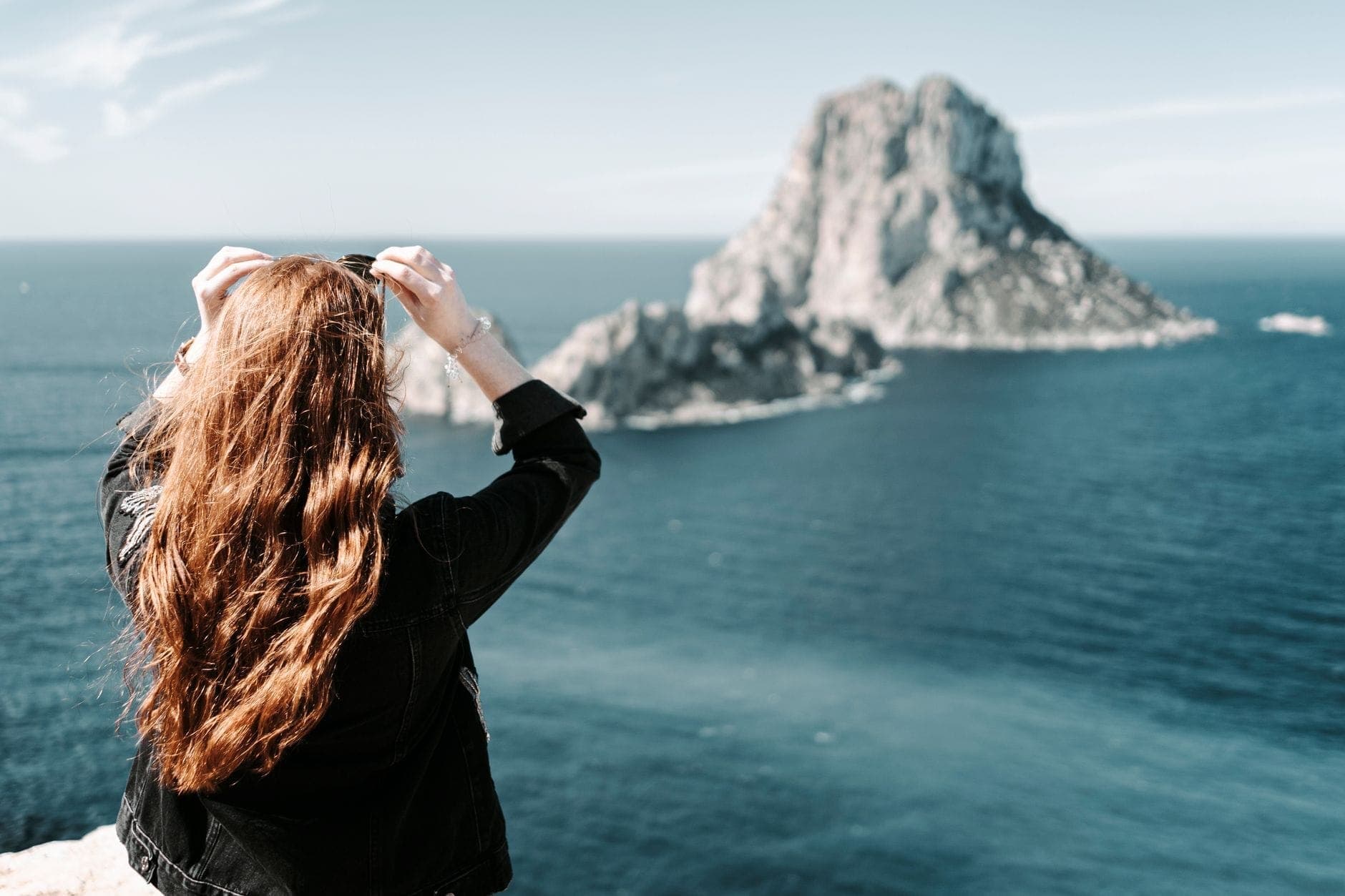 A woman with long hair stands overlooking the blue sea and Es Vedrà island off the coast of Ibiza on a sunny day.