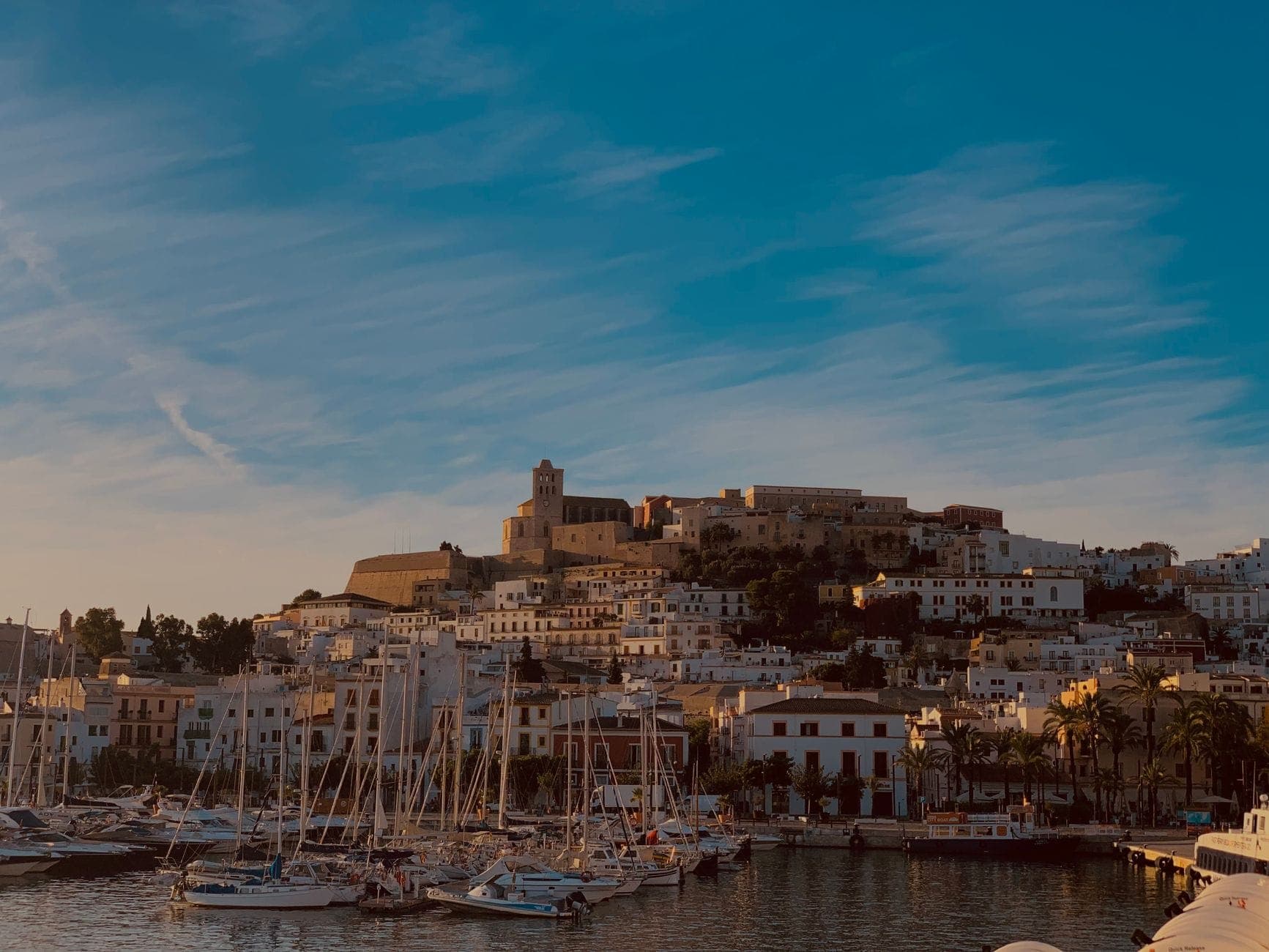 View of Ibiza old town and harbor at sunset, with white buildings, ancient fortress, and boats docked in the marina under a blue sky.