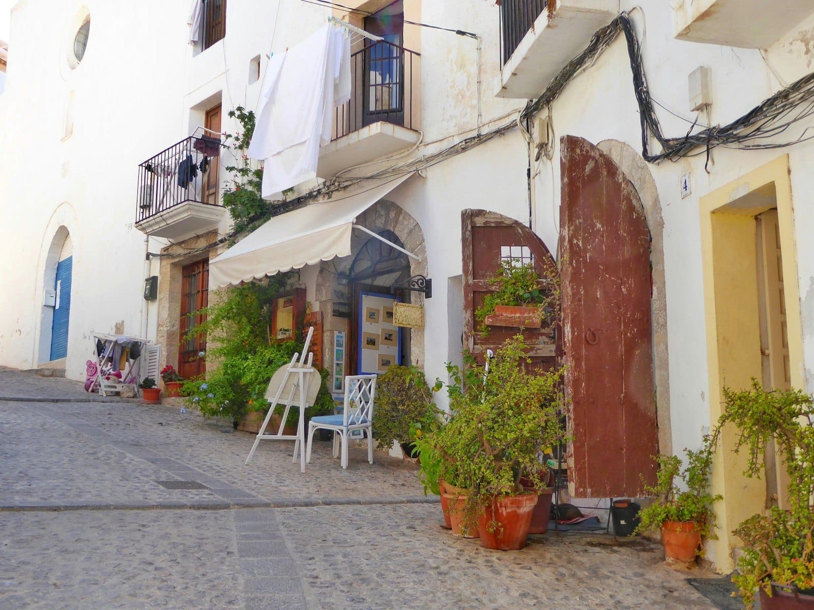 Cobbled street in Ibiza's Dalt Vila old town with whitewashed buildings, potted plants, wooden doors, and laundry hanging from balconies.