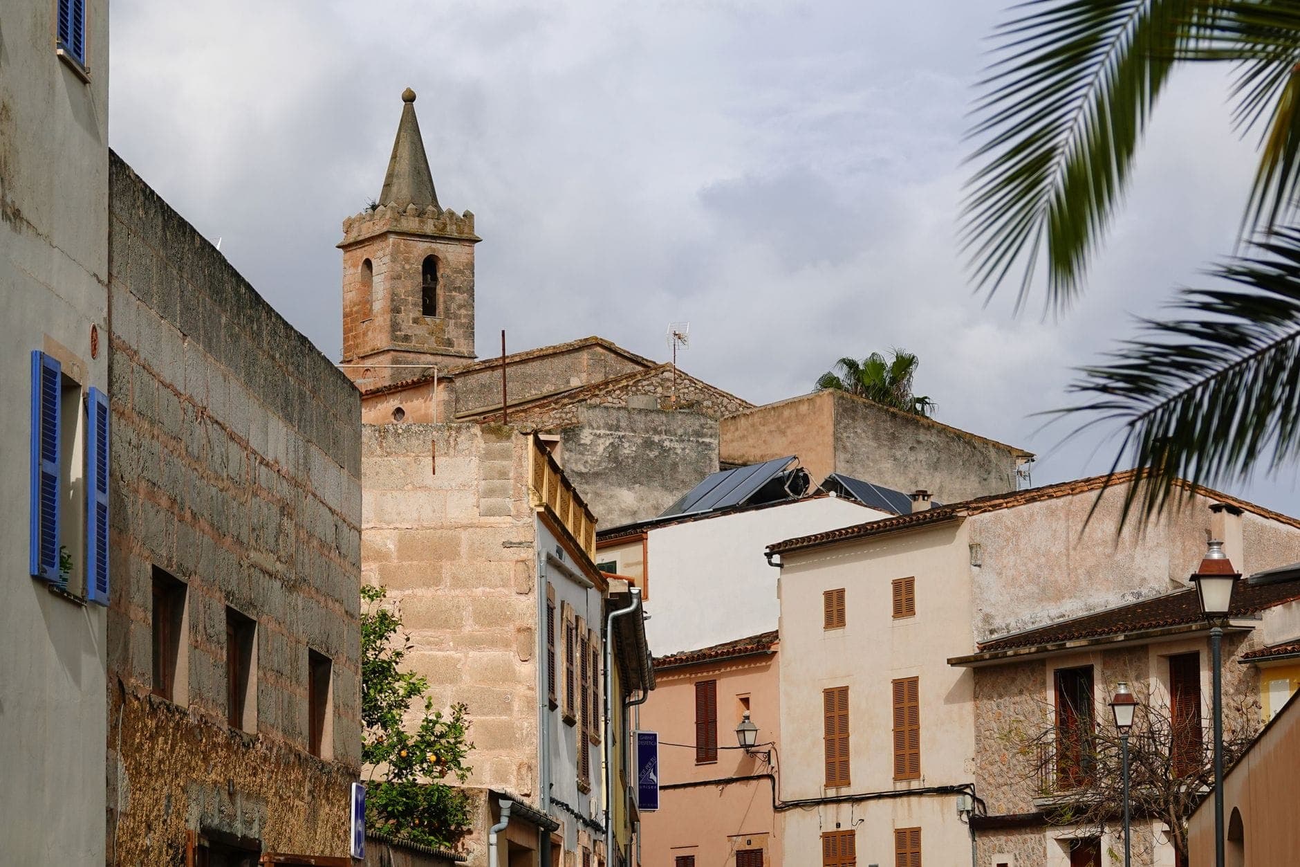 View of a Mediterranean village with whitewashed buildings and a church tower, surrounded by palm trees and narrow streets.