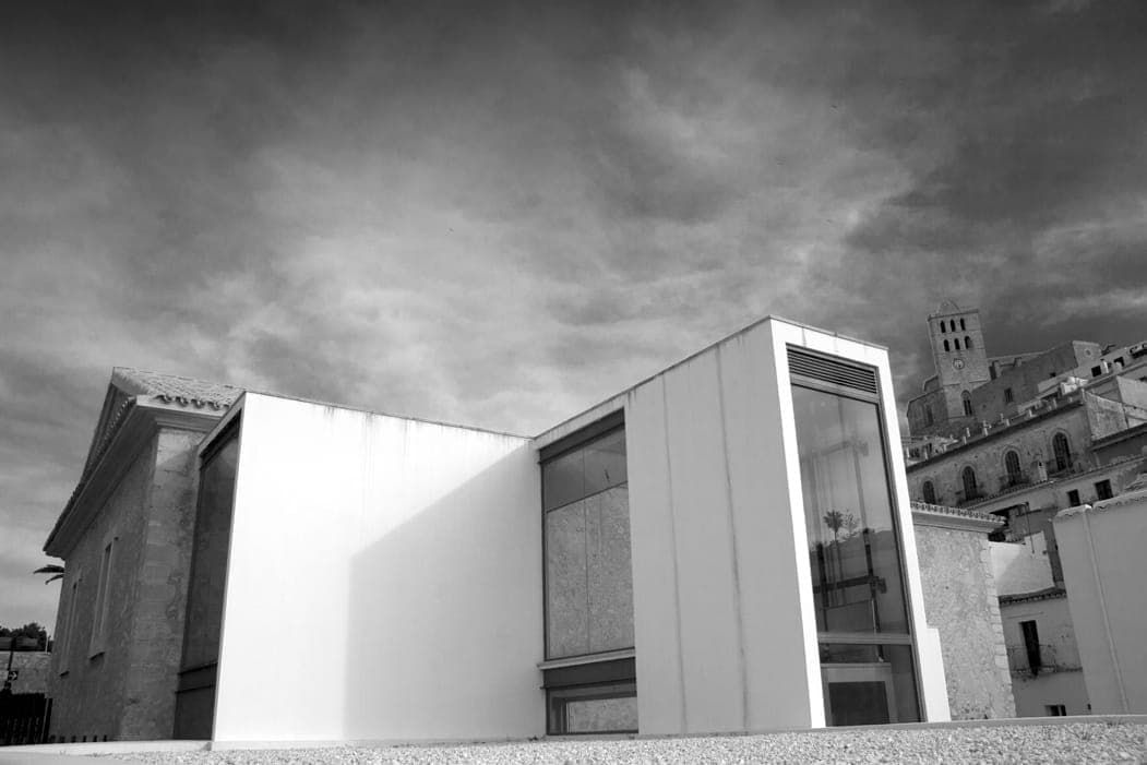 Exterior view of Ibiza Museum of Contemporary Art (MACE) featuring modern glass architecture beside historic stone buildings under a dramatic cloudy sky.