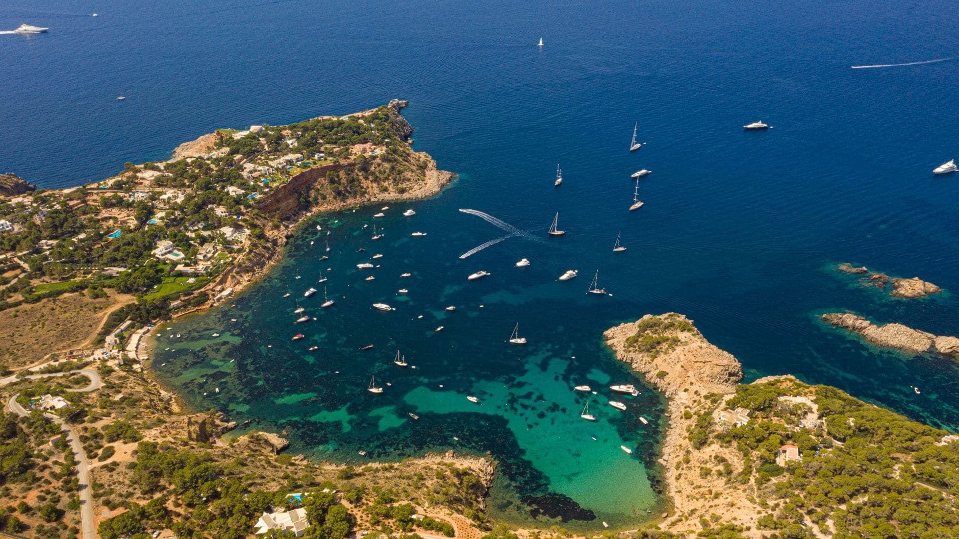 Aerial view of a bay in Ibiza with numerous boats and yachts anchored in clear turquoise water, surrounded by rocky coast and green hills.