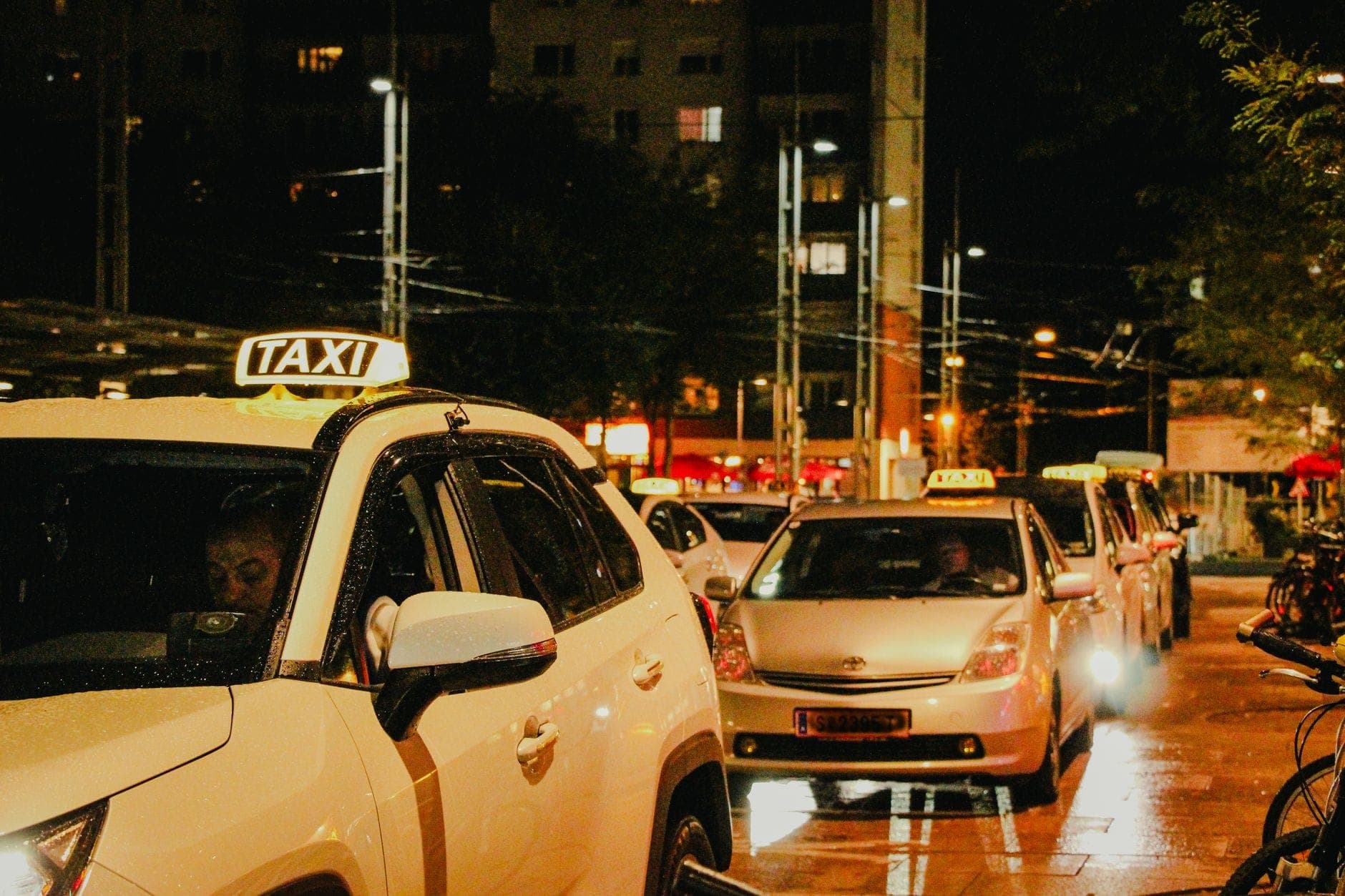 A line of white taxis waiting at night on a wet, well-lit city street with buildings and streetlights visible in the background.