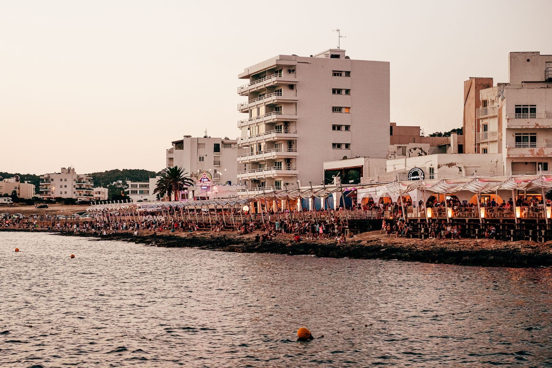 Lively row of seaside bars and restaurants crowded with people at dusk, with modern buildings and palm trees in the background, capturing Ibiza nightlife atmosphere.