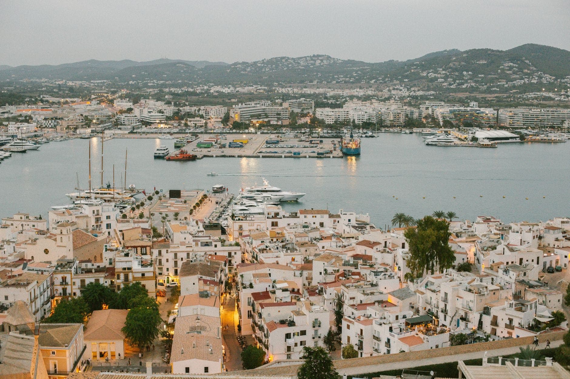 Wide view of Ibiza town and marina with yachts in harbor, white buildings, and hills in the distance at dusk.
