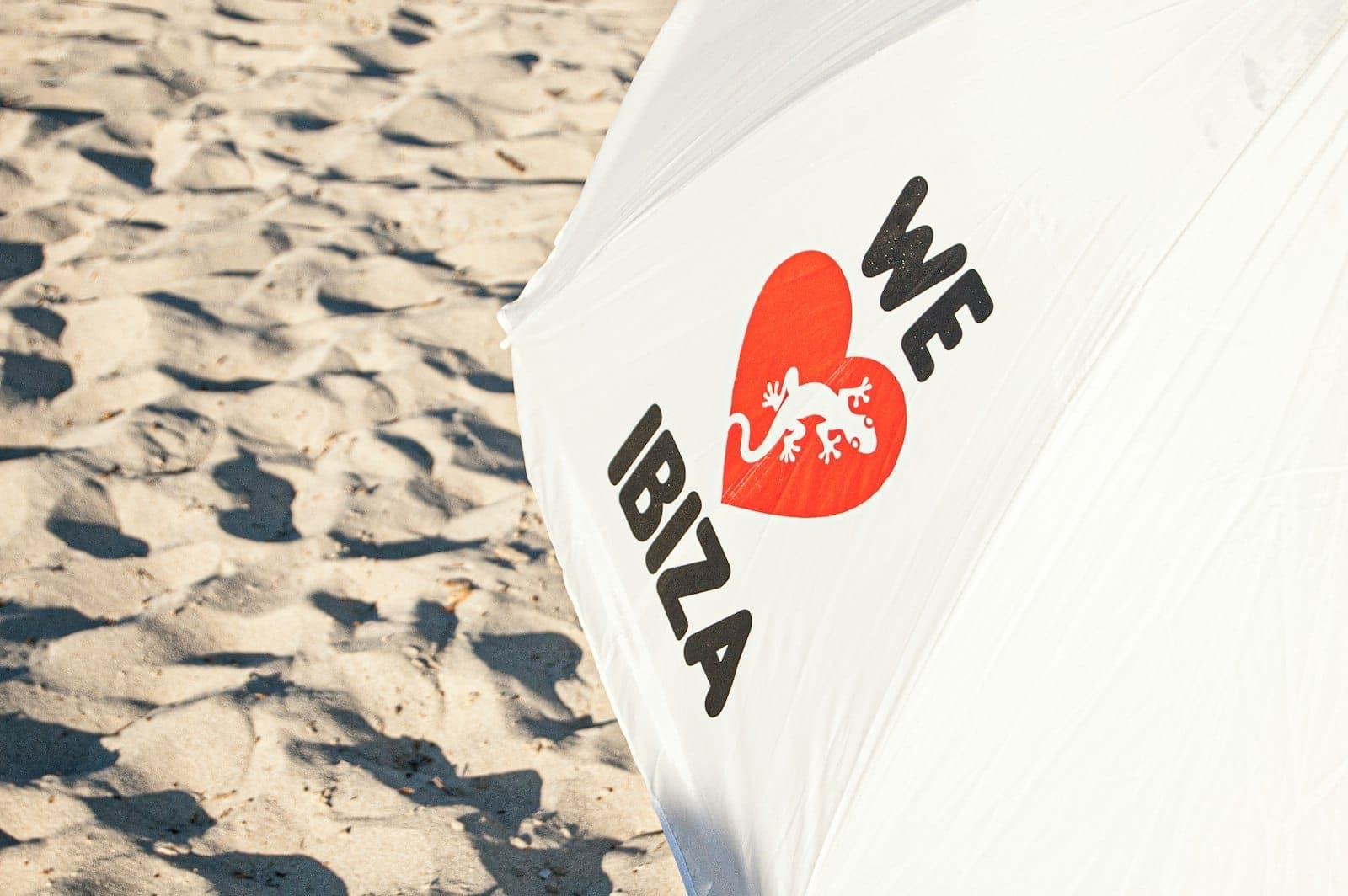 Close-up of a white beach umbrella with 'We Love Ibiza' text and a red heart, set against sandy beach background.