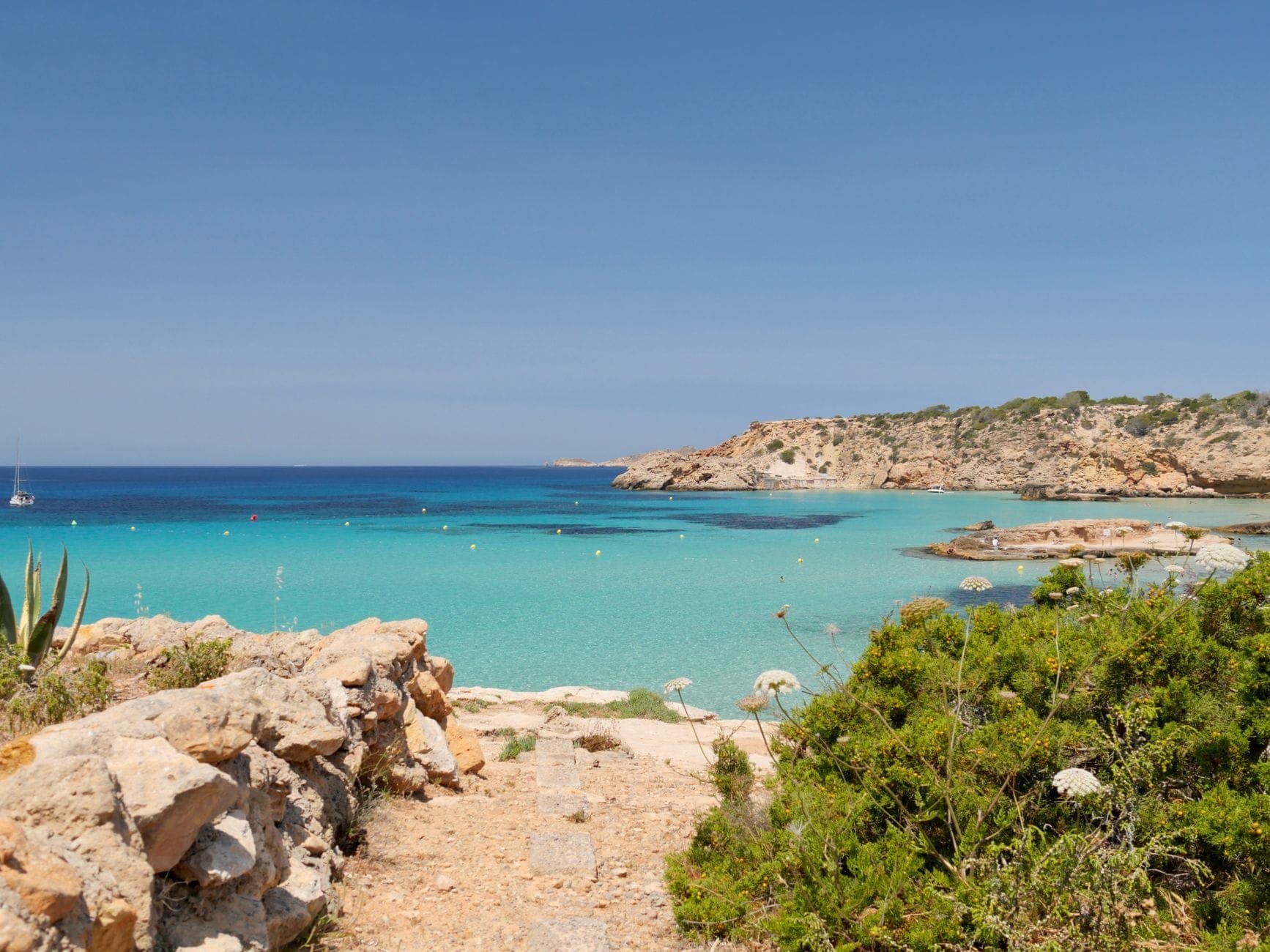 Sunny coastal scene in Ibiza with turquoise water, rocky shoreline, and clear blue sky, showcasing a free public beach area.