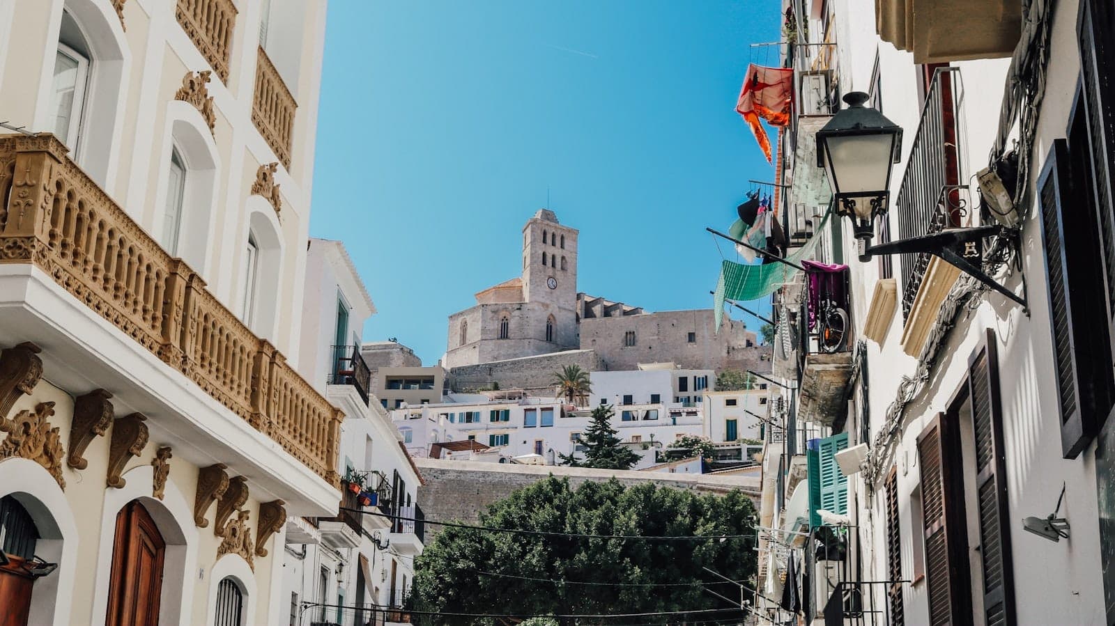 View up a charming street in Ibiza Town with traditional white buildings, balconies, and the historic Dalt Vila fortress above under a clear blue sky.
