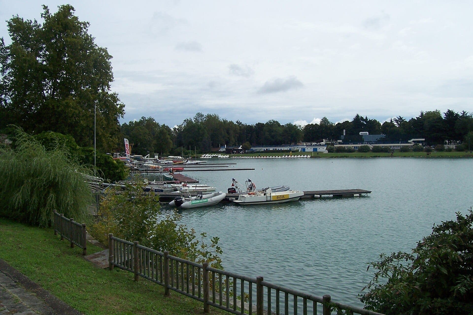 Boats moored at a wooden dock along the tree-lined artificial lake at Idroscalo di Milano under a cloudy sky.