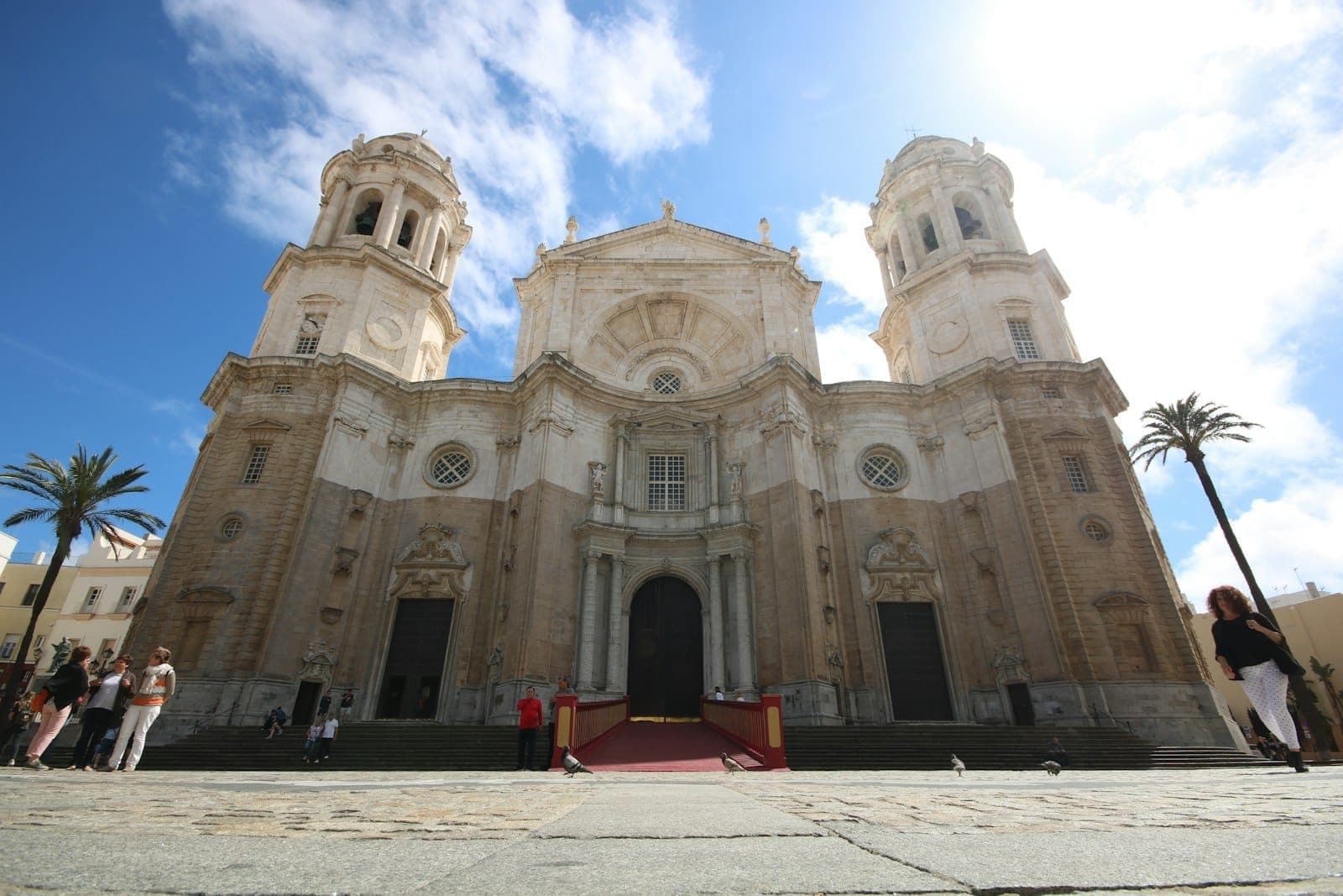 Nhìn từ phía trước Iglesia de Santa Cruz ở Cádiz, với hai tháp chuông đôi, mặt tiền bằng đá chạm khắc tinh xảo và người dân tụ tập tại quảng trường ngập nắng.