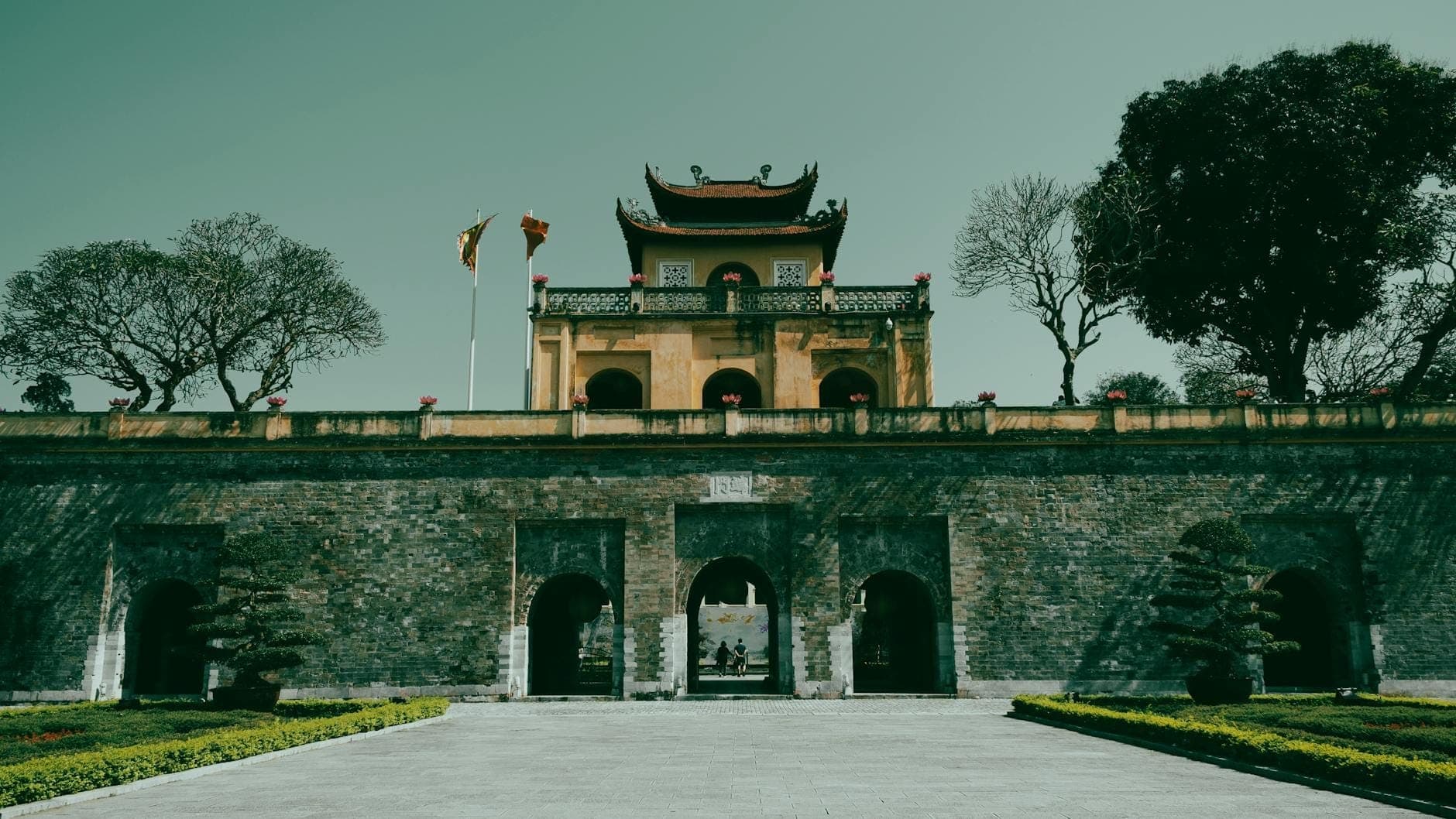 Toàn cảnh cổng chính và tòa nhà trung tâm của Hoàng thành Thăng Long tại Hà Nội, hai bên có cờ và cây xanh dưới bầu trời trong xanh.