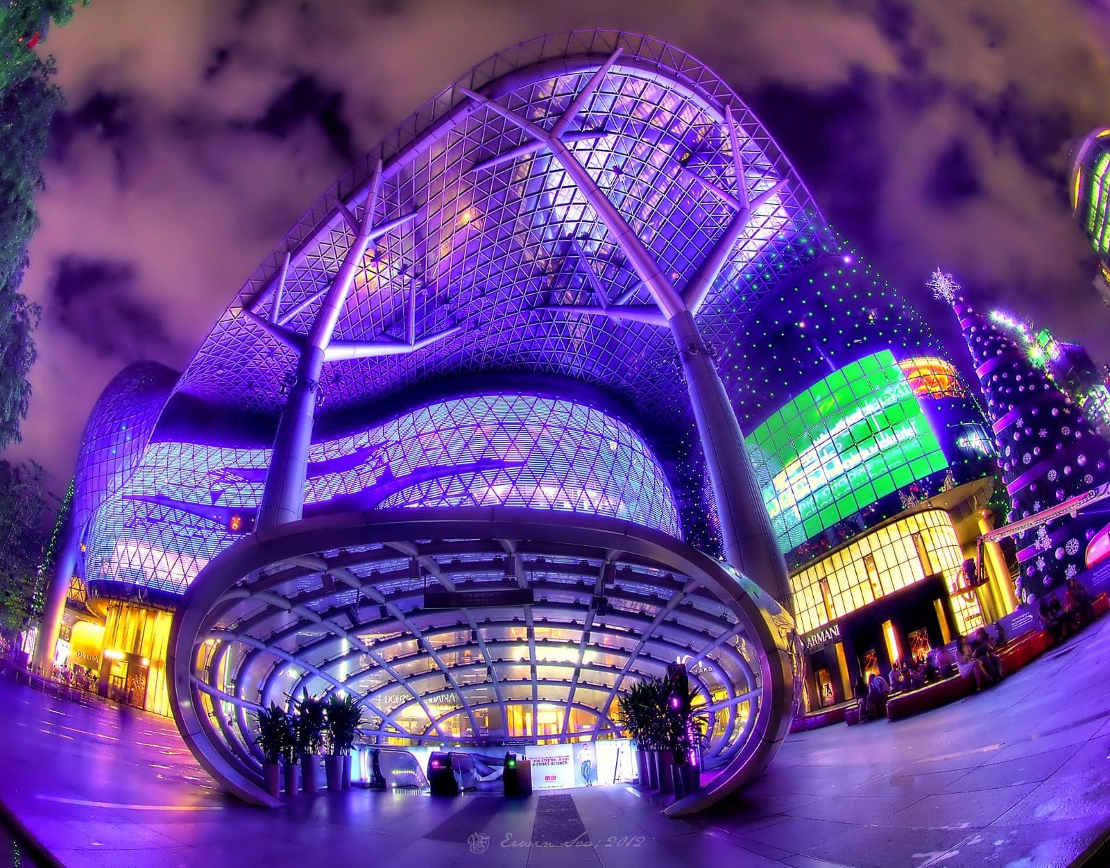 Wide-angle view of ION Orchard’s futuristic glass and steel exterior illuminated with purple, blue, and green lights at night, attracting visitors on Orchard Road.