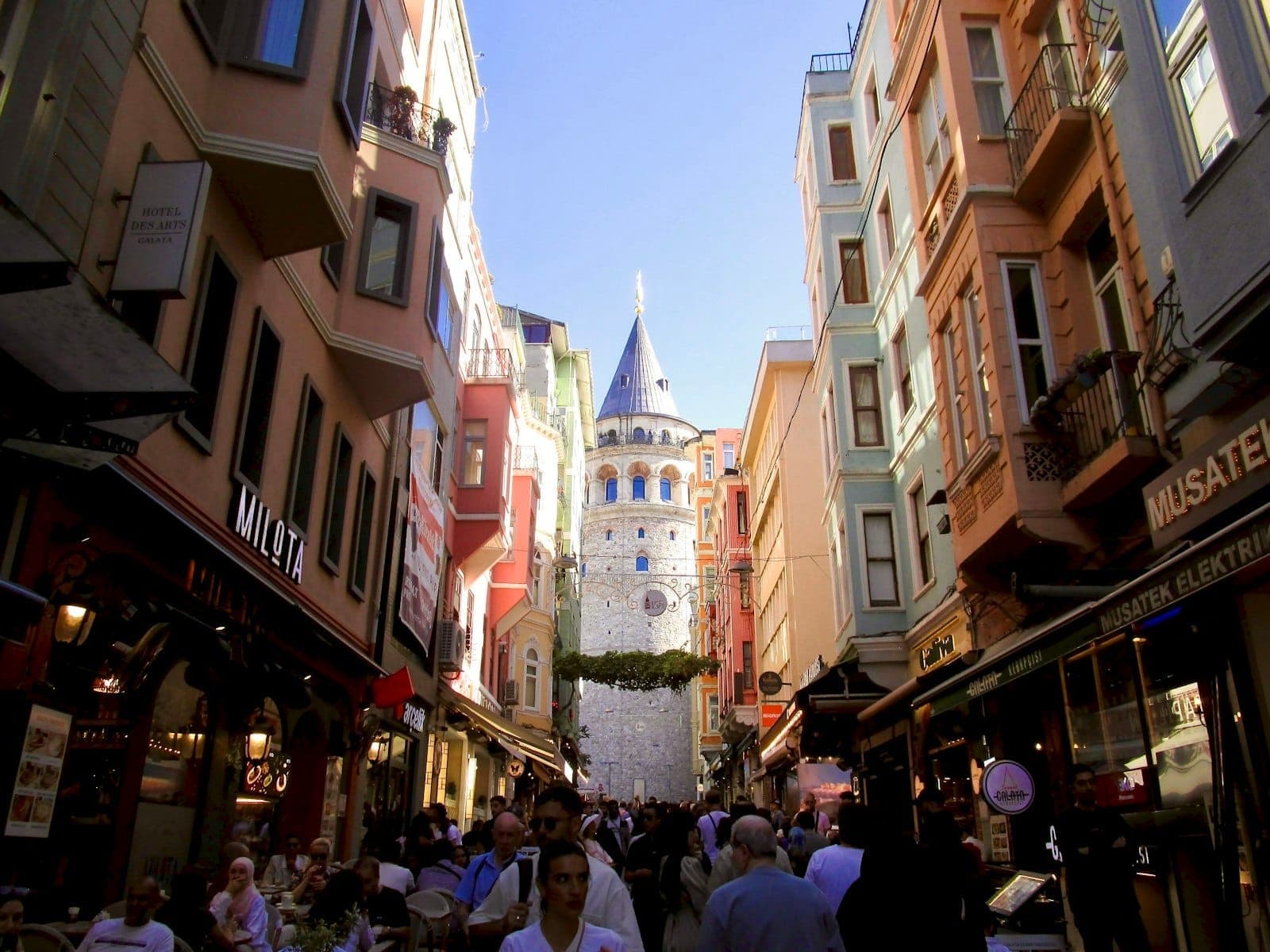 Jalan pejalan kaki yang ramai di Istanbul dikelilingi gedung-gedung berwarna-warni, kafe, dan Menara Galata terlihat di kejauhan di bawah langit cerah.